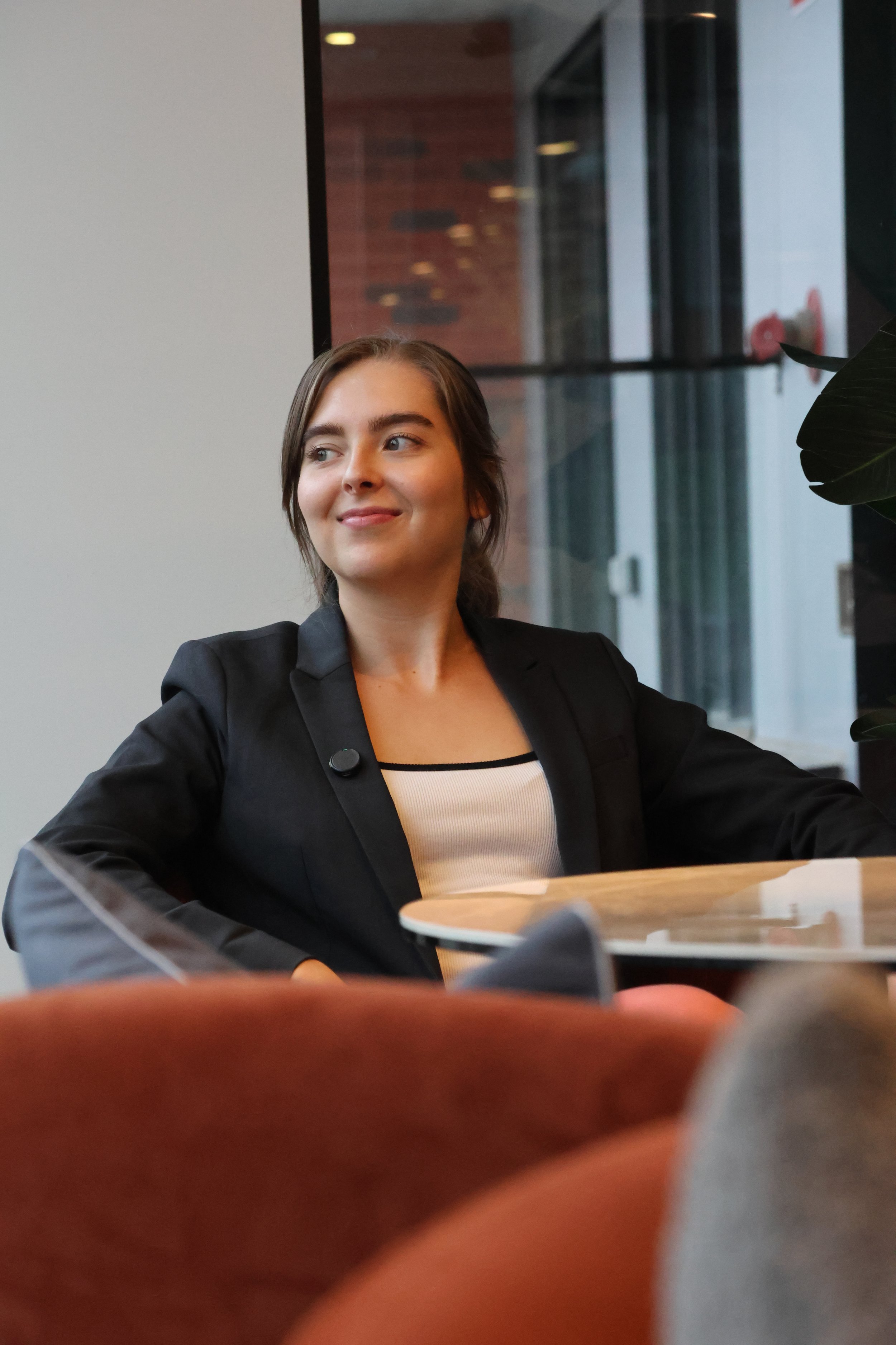 A young woman with long brown hair, wearing a black blazer and white top, sitting at a table in a modern indoor setting with a large window in the background.