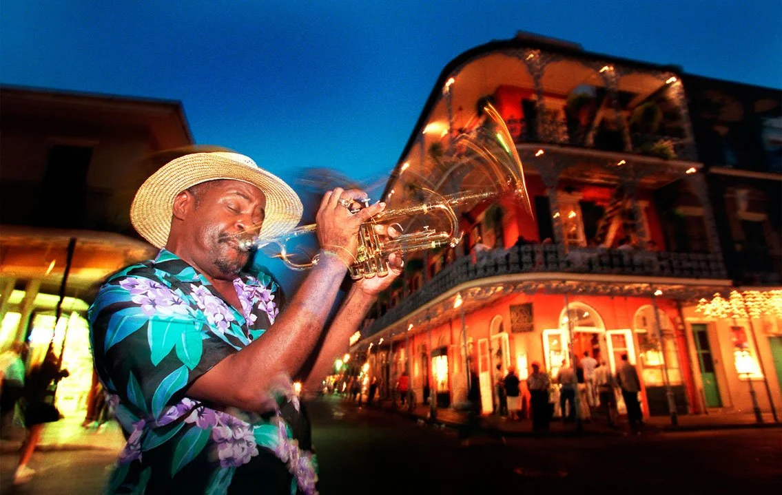 A man wearing a floral shirt and straw hat playing a trumpet on a city street at dusk, with a brightly lit building in the background.