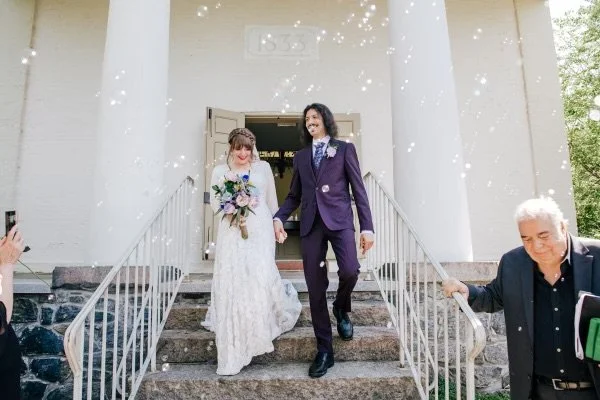 A bride and groom exiting a building after a wedding, holding hands and smiling, with confetti falling around them.