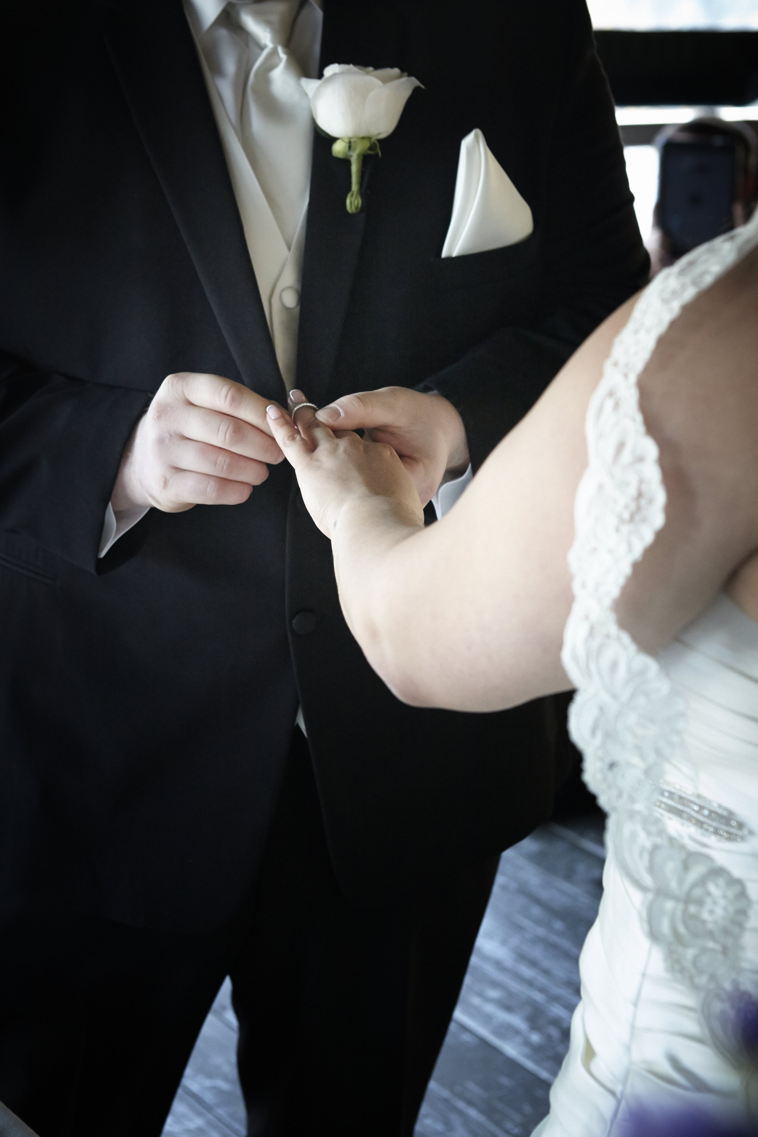 A groom in a black tuxedo and a white boutonniere holds the bride's hand as he places a wedding ring on her finger during their wedding ceremony.