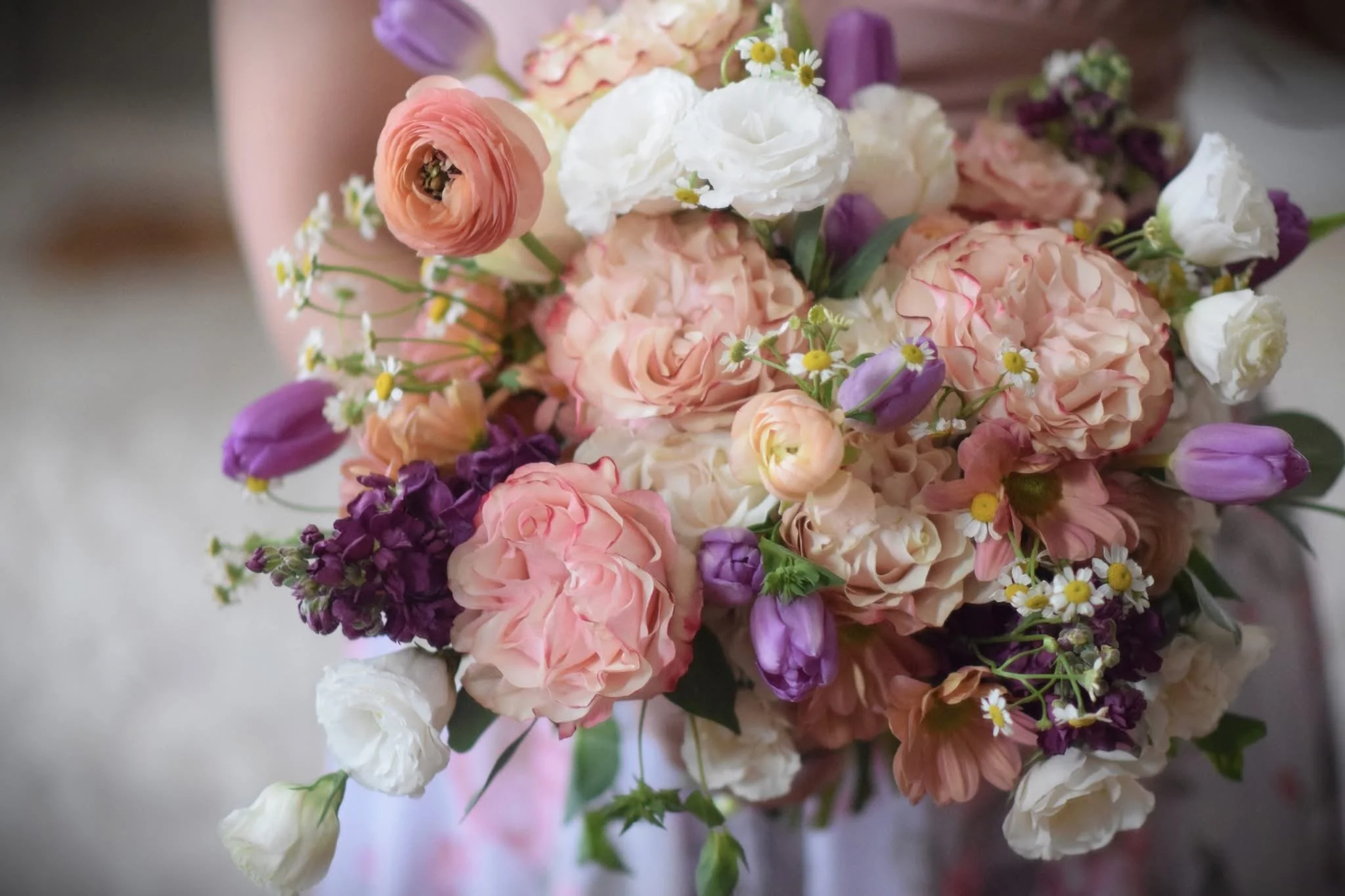 A bride holding a cascading bouquet of peach roses, white anemones with dark centers, cream-colored ranunculus, and greenery.