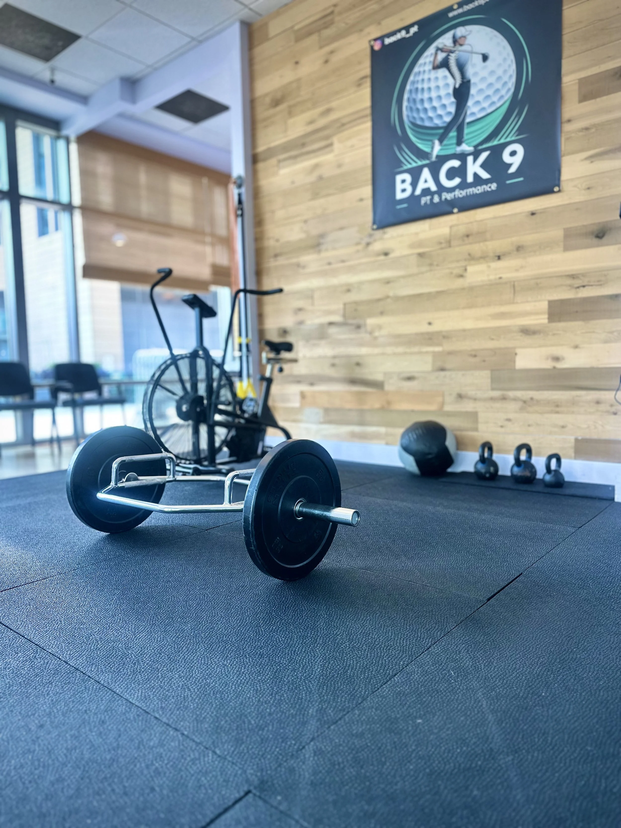A professional photo of the interior of the Back 9 PT & Performance clinic in Cambridge, MA, showing a variety of exercise equipment including an assault bike, kettlebells, and a hex bar.
