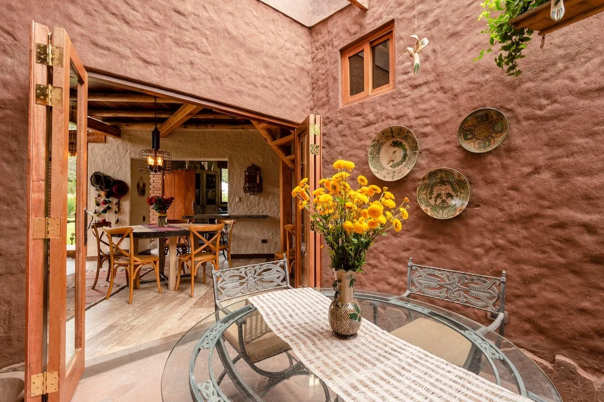 A cozy indoor-outdoor dining area with a glass table, a vase of yellow flowers, and decorative plates on a pink stucco wall.