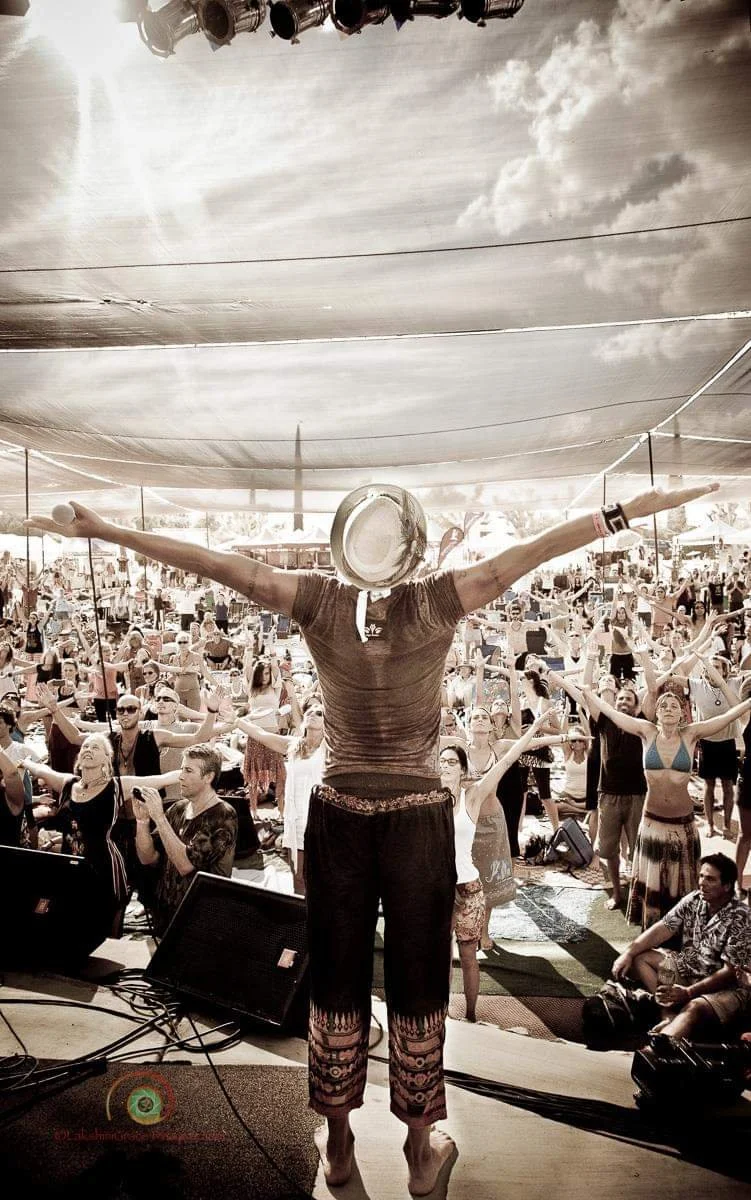 Person with a hat on stage leading a large crowd at a festival or concert under a canopy.