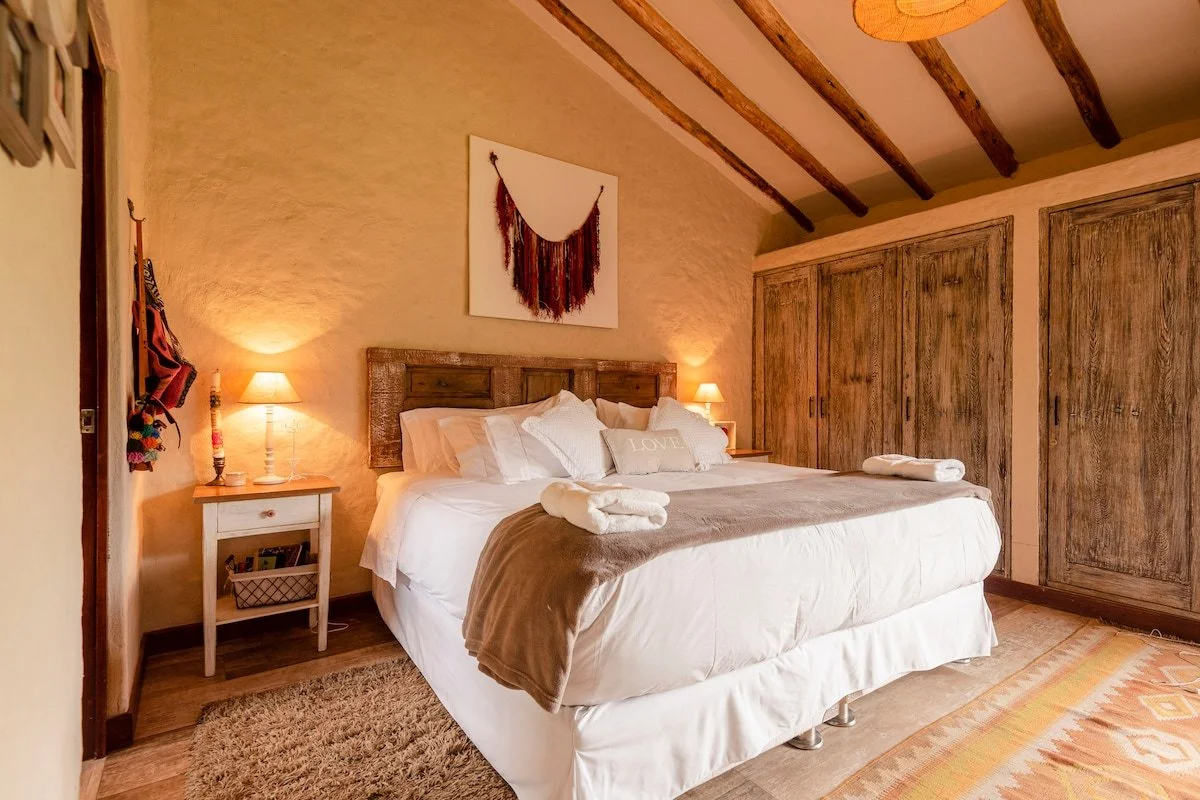 Cozy bedroom with a large bed, wooden headboard, white bedding, decorative pillows, and folded towels. Two small bedside lamps, wooden beams on the ceiling, closet with wooden doors, and artwork hanging above the bed.