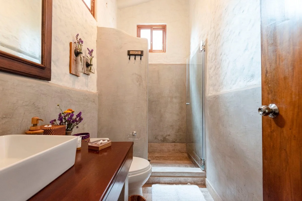 A small bathroom with a wooden door, a sink with a wooden countertop, decorated with soap and flowers, a toilet, a walk-in shower with a glass divider, a small window above, and light-colored textured walls.