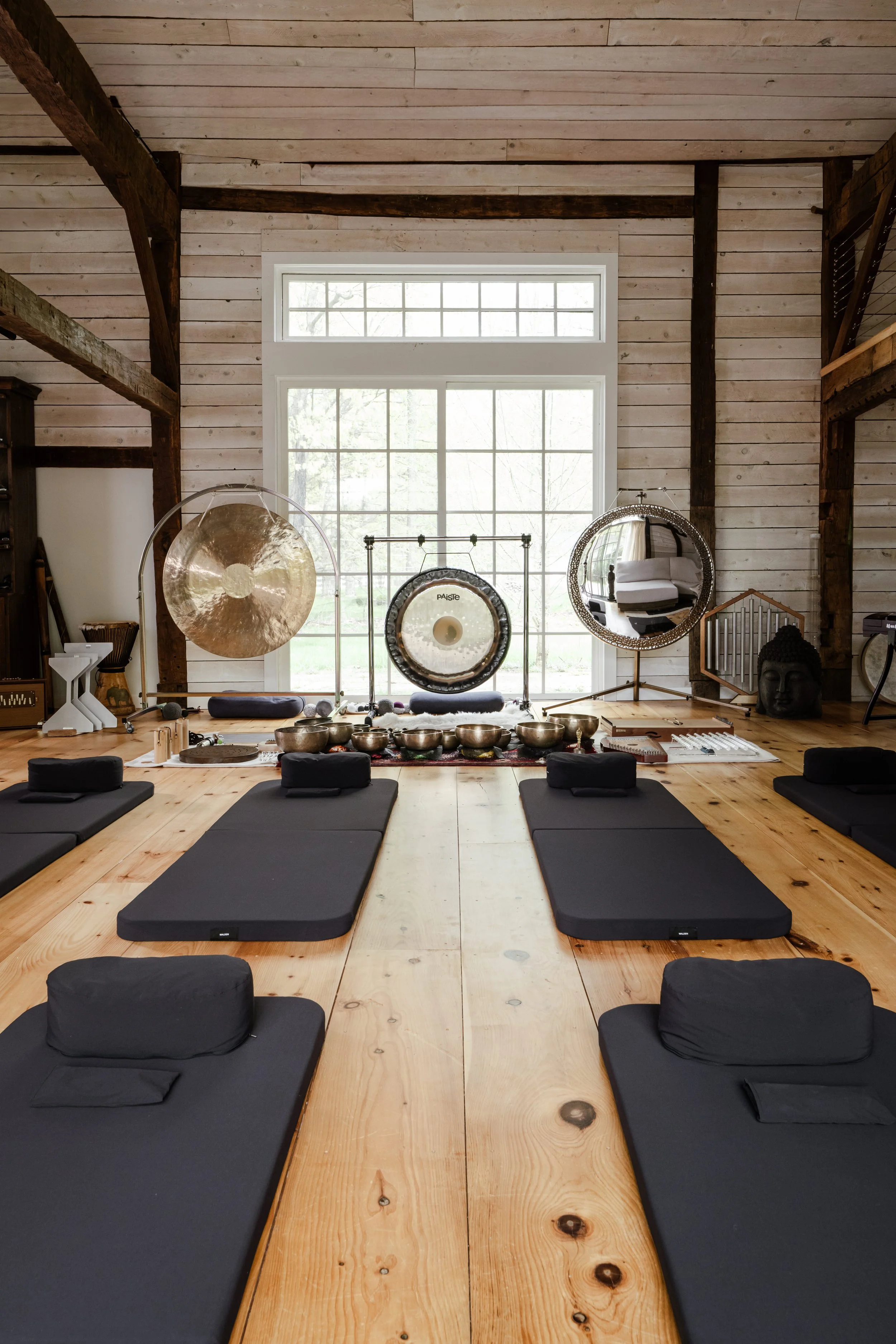 A tranquil yoga or meditation space with black mats and cushions arranged on a wooden floor, facing a large window that lets in natural light. The room features white and dark wood paneling with visible wooden beams, and decorative meditation gongs and other spiritual ornaments are placed along the back wall.