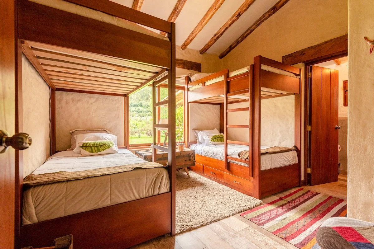 A bedroom with three wooden bunk beds, a window showing greenery outside, and a striped rug on the floor.