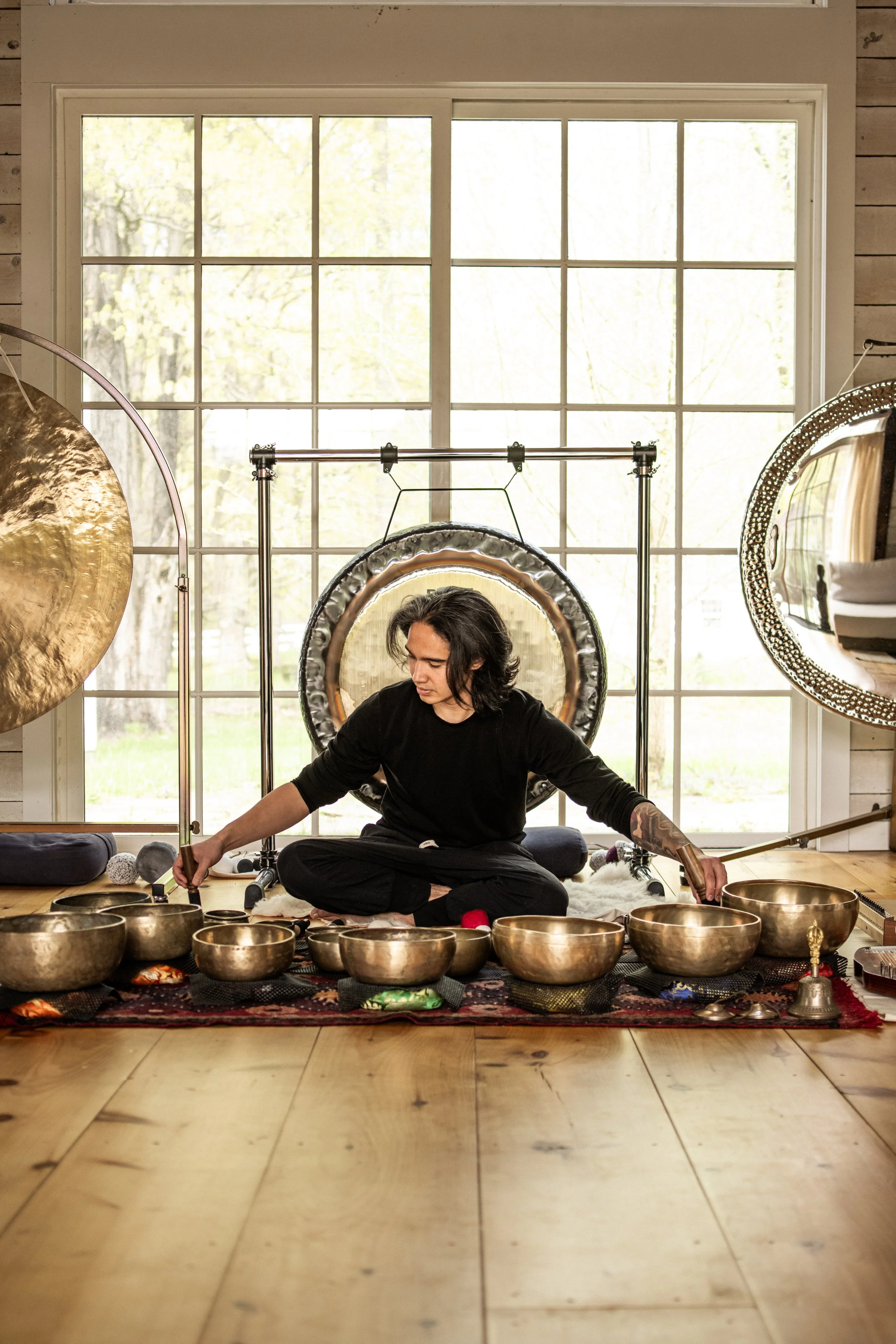A person with long dark hair, wearing a black shirt, sitting cross-legged on a patterned rug inside a room with large windows, surrounded by metallic singing bowls of various sizes, engaging in a sound therapy session.