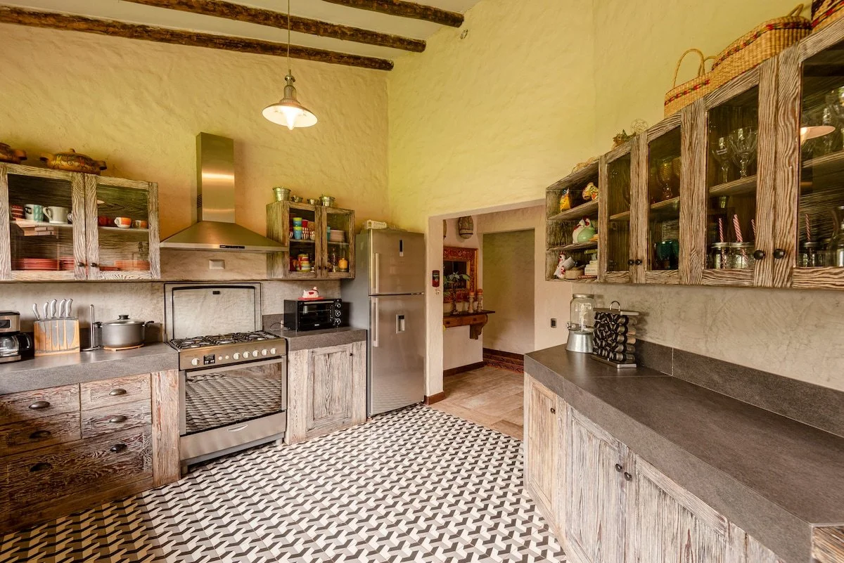 Rustic kitchen with wooden cabinets, a stainless steel refrigerator, a stove, and open shelving with dishes and glassware. Patterned tile floor and exposed wooden ceiling beams.