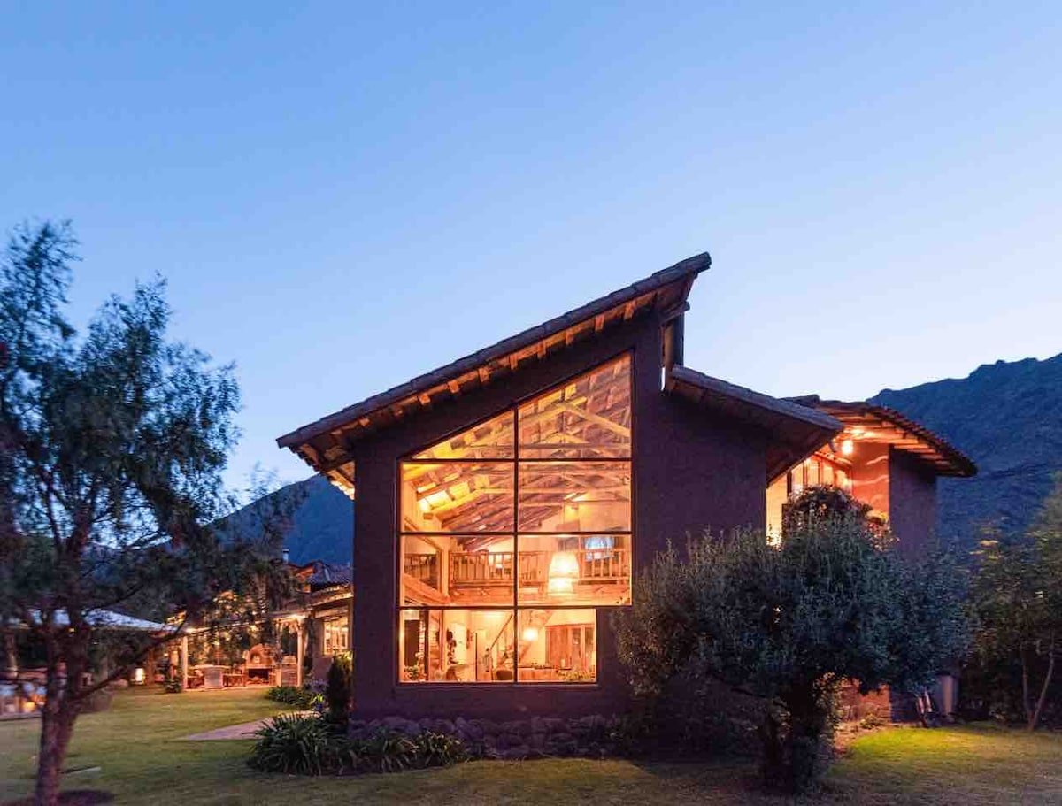A modern house with large glass windows glowing with warm interior lights, surrounded by a lawn and trees, with mountains in the background at dusk.