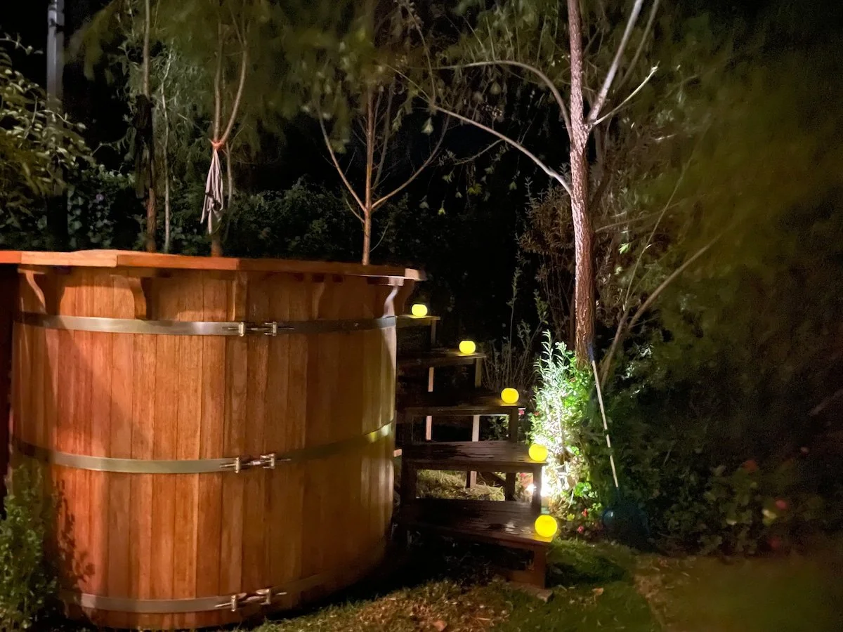Nighttime scene featuring a wooden hot tub on the left and a small staircase decorated with yellow glowing orbs on the right, surrounded by trees and bushes.