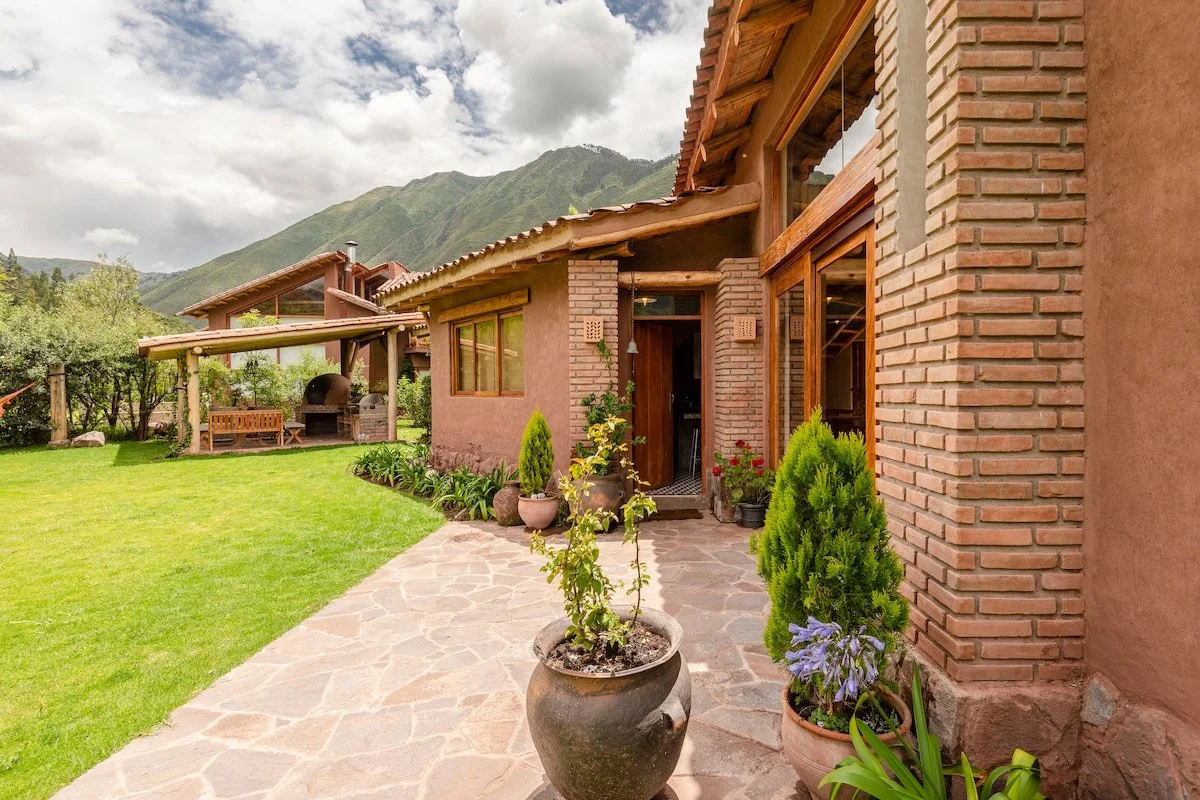 Exterior view of a house with a brick and stucco facade, large windows, a stone pathway, and potted plants, with green mountains in the background.
