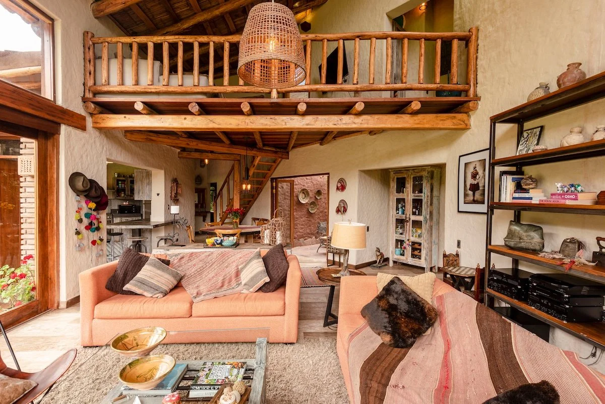 Living room with two pink sofas, decorative pillows, and a wooden coffee table, featuring a rustic style with a mezzanine and a staircase, shelves with books and ornaments, and a small table lamp.