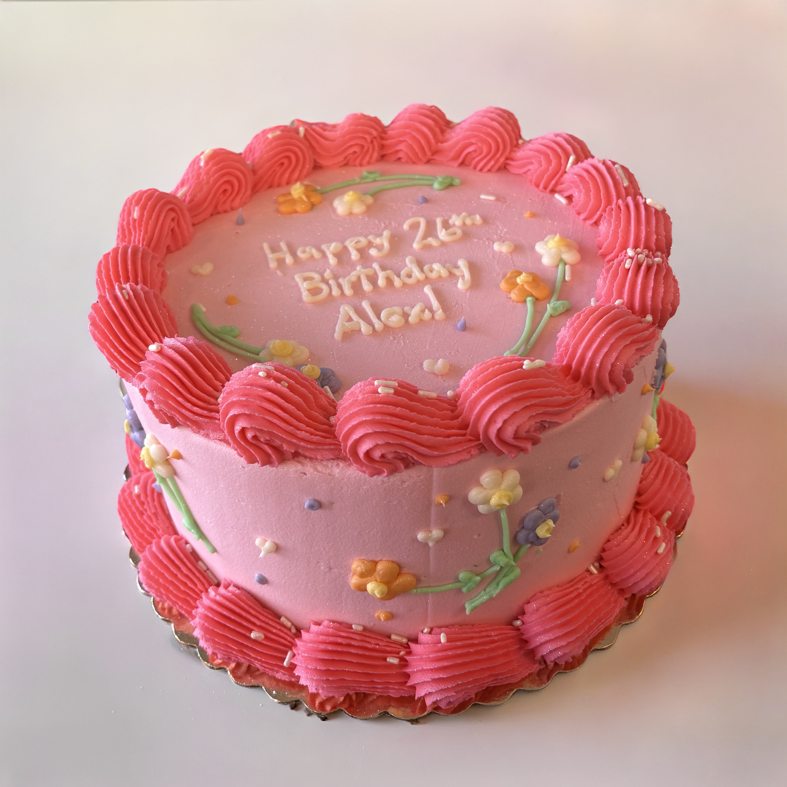 Pink birthday cake decorated with pink frosting swirls, colorful flower sprinkles, and a message that reads "Happy 26th Birthday Alec" in the center.