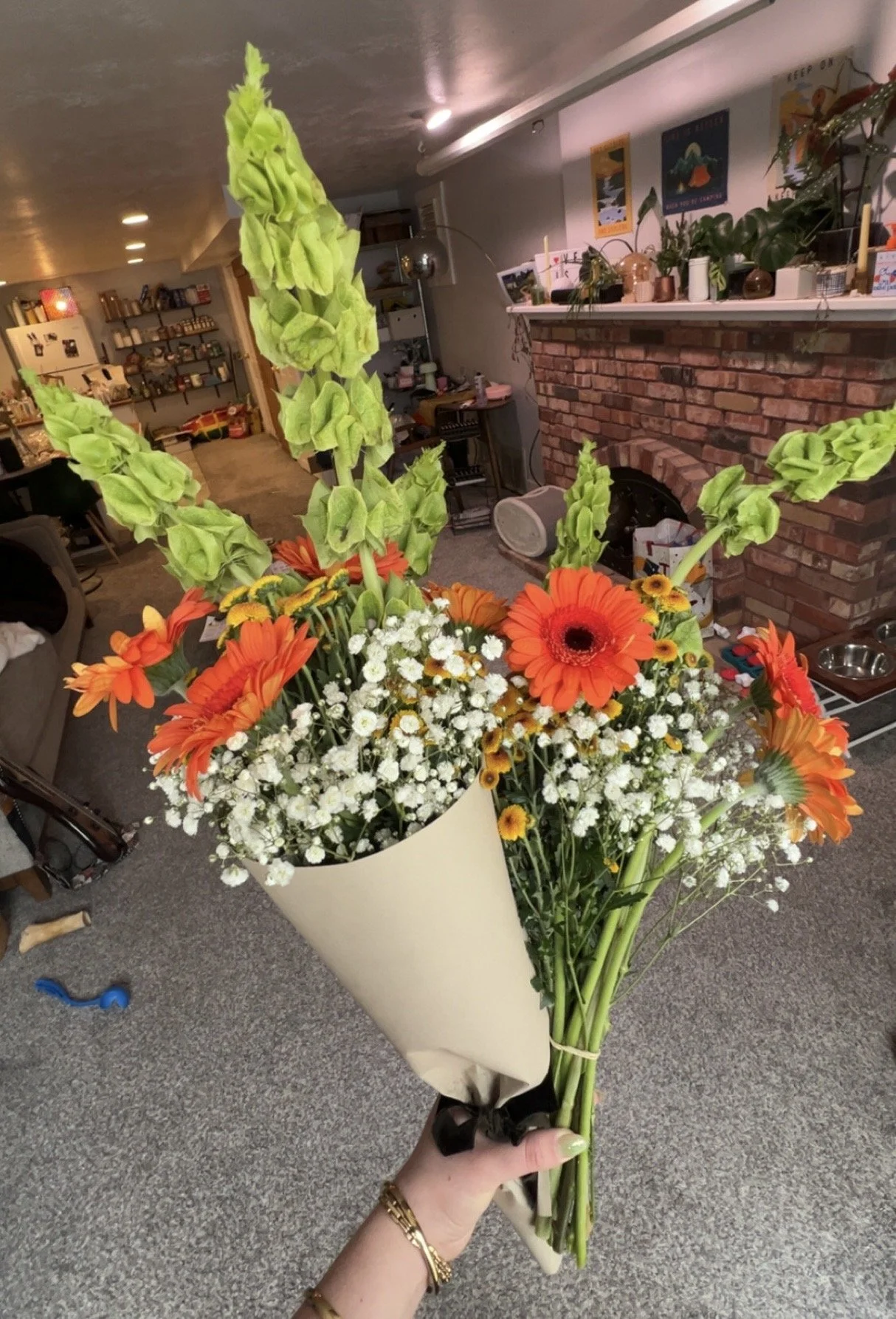 A hand holding a bouquet of flowers with bright orange gerbera daisies, white baby's breath, and tall green snapdragons, wrapped in beige paper with a black ribbon, in a cozy indoor living space with a brick fireplace and bookshelves.