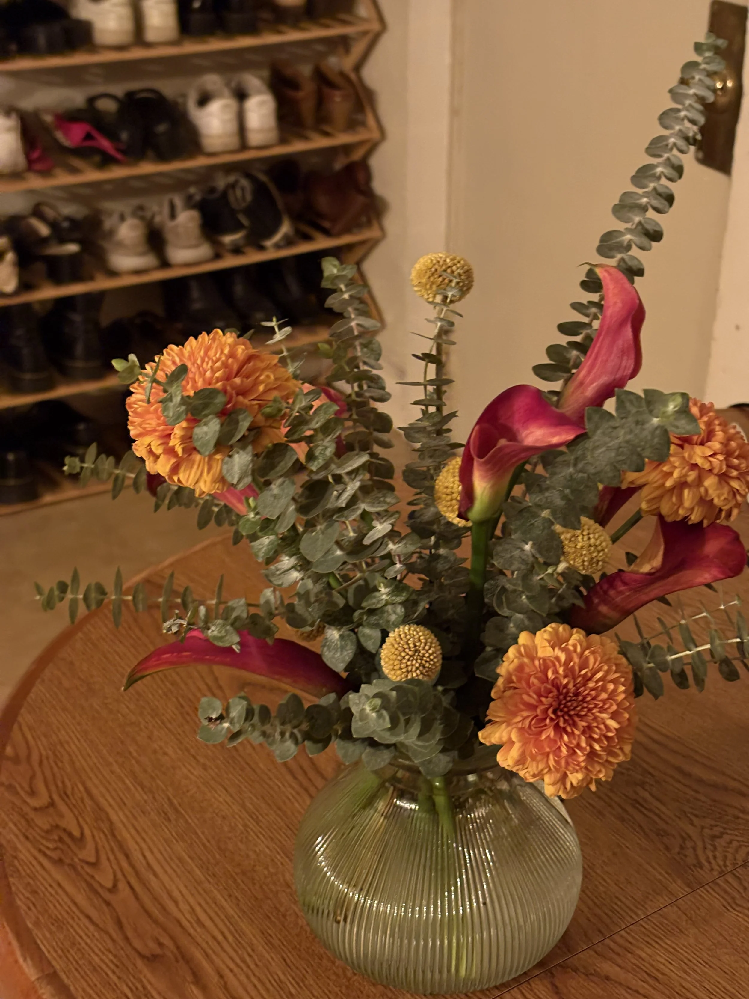 A glass vase holding a floral arrangement with orange and red flowers, yellow round flowers, and eucalyptus leaves on a wooden table.
