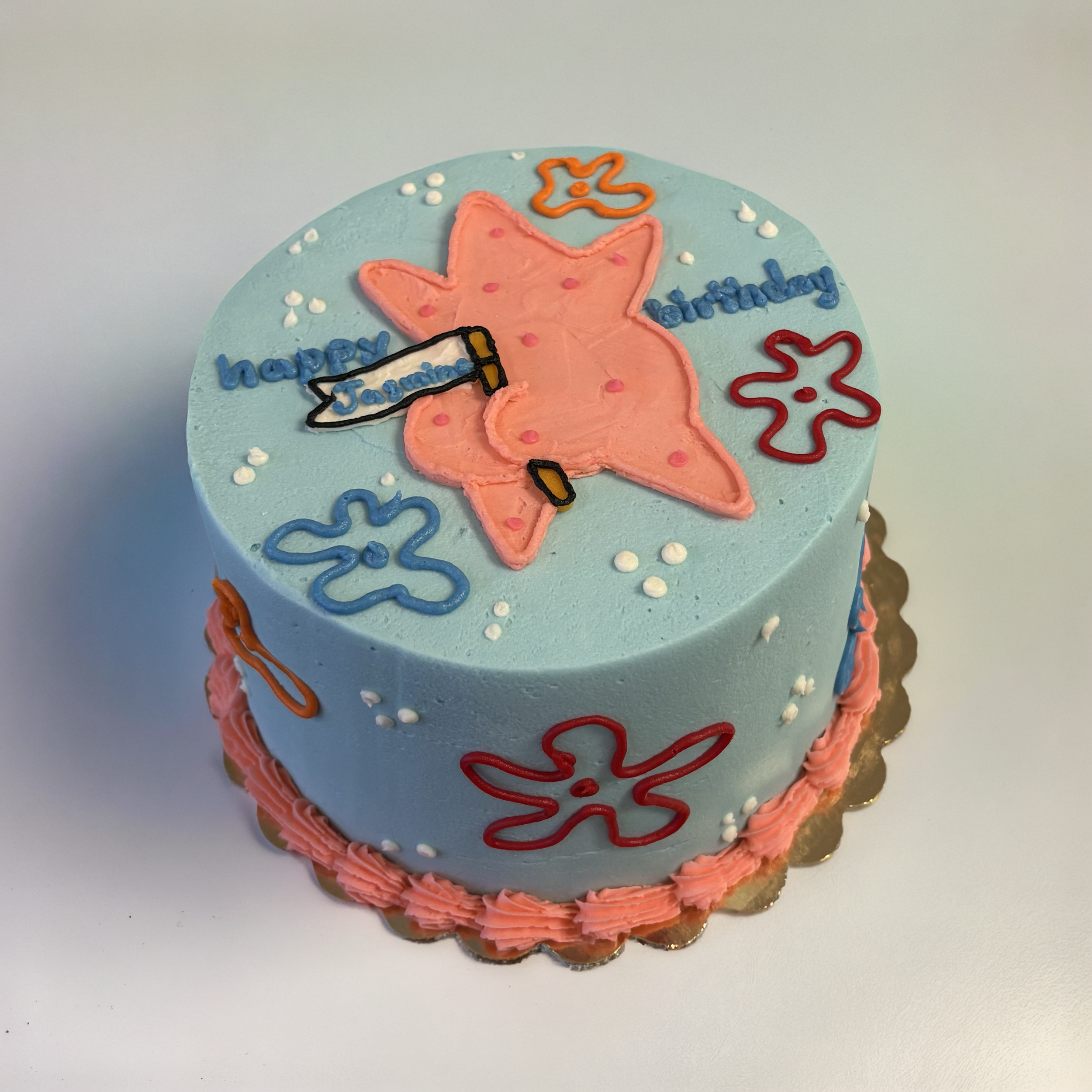A round birthday cake with pink, orange, blue, and white decorations including a starfish, flowers, and bubbles. The cake has a pink border at the bottom and a message that says 'Happy Birthday' with a starfish and a hammer and sickle on top.