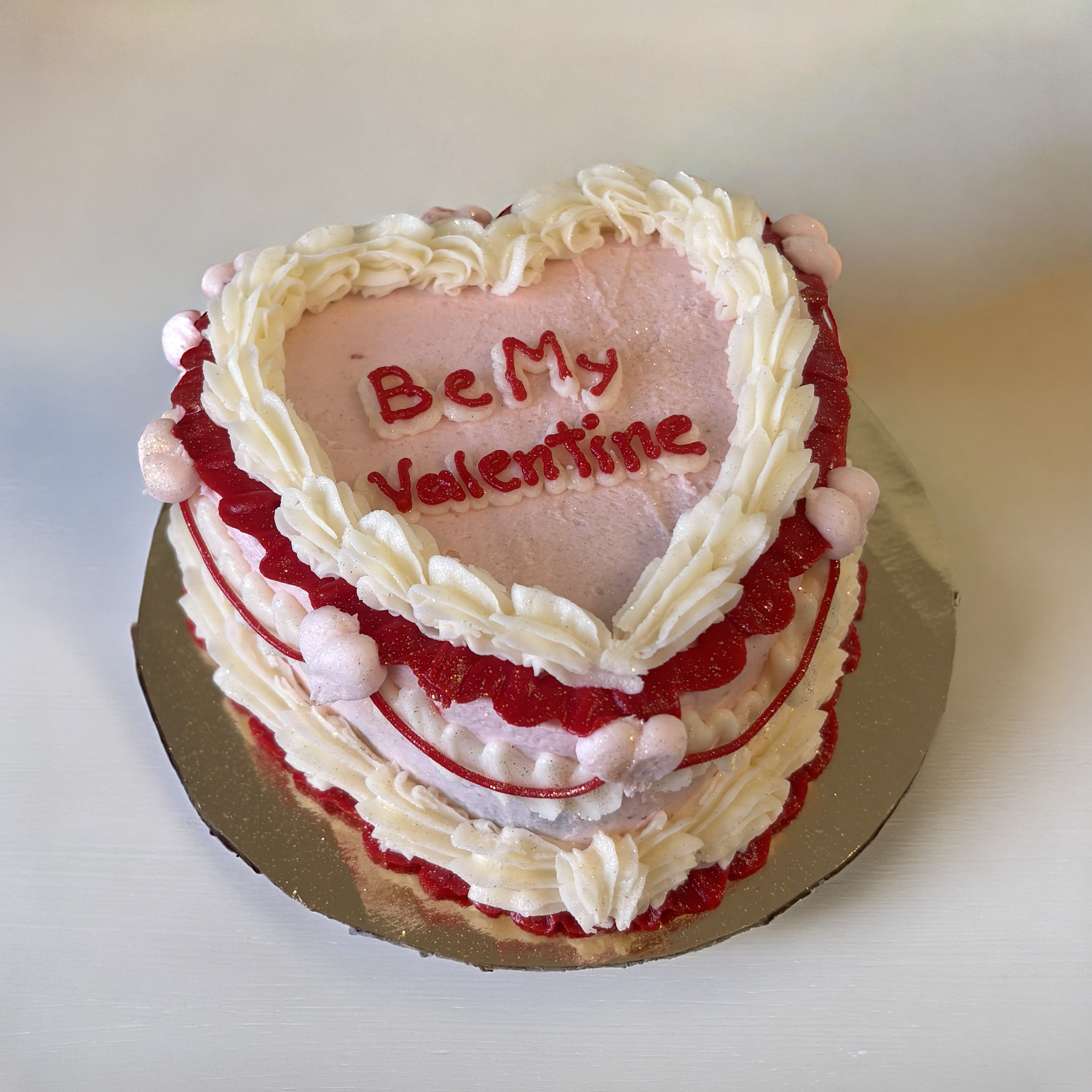 Heart-shaped Valentine's Day cake with white and pink frosting, red accents, and the message "Be My Valentine" written on top.