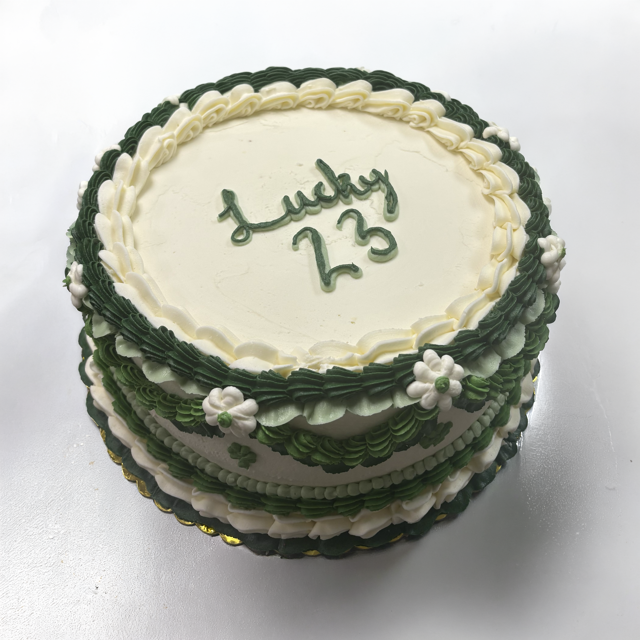 A round birthday cake with green and white icing decorations, and a birthday message written in green icing on top, placed on a white surface.