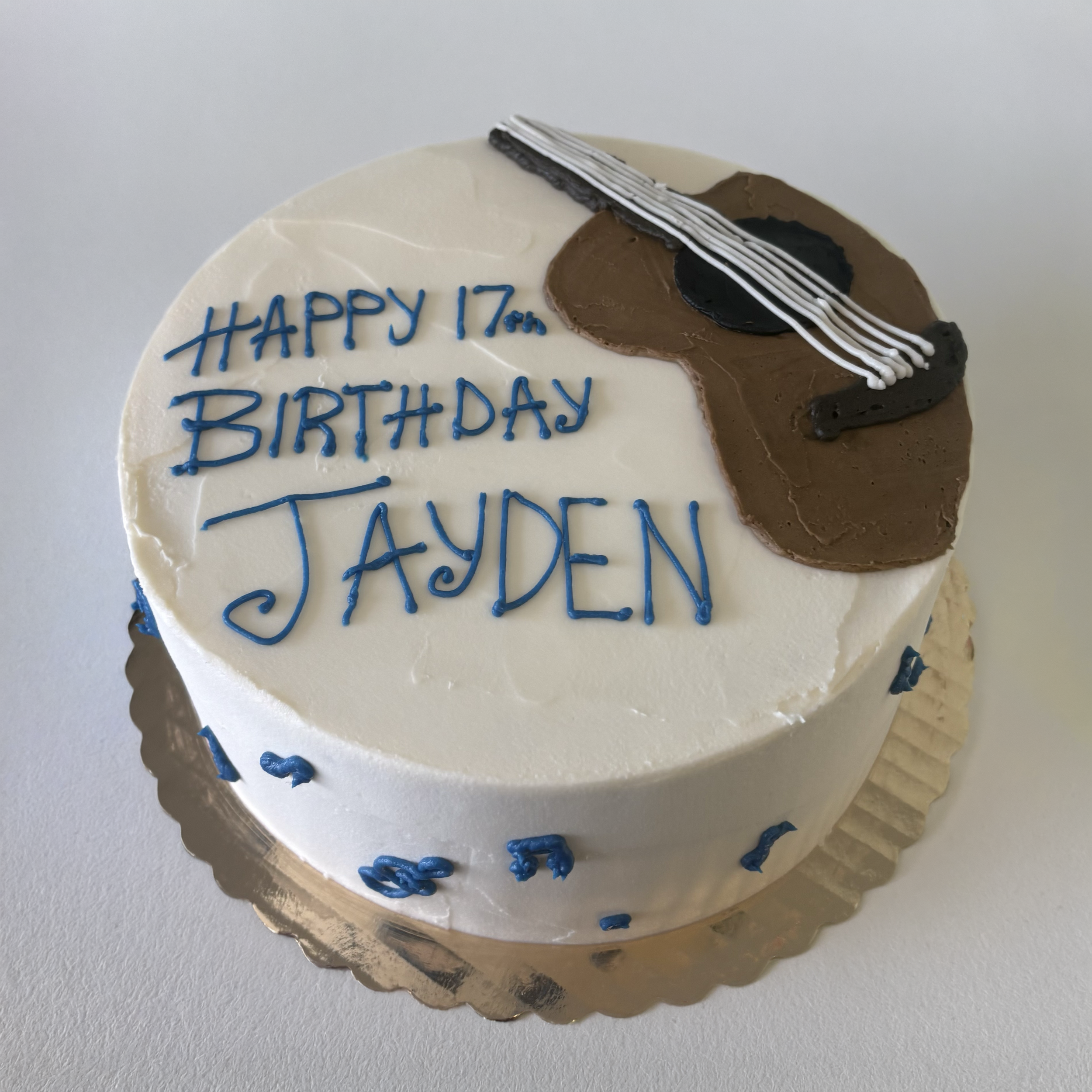 A round birthday cake with white frosting, decorated with a guitar design and a message that reads 'Happy 17th Birthday Jayden' written in blue icing.
