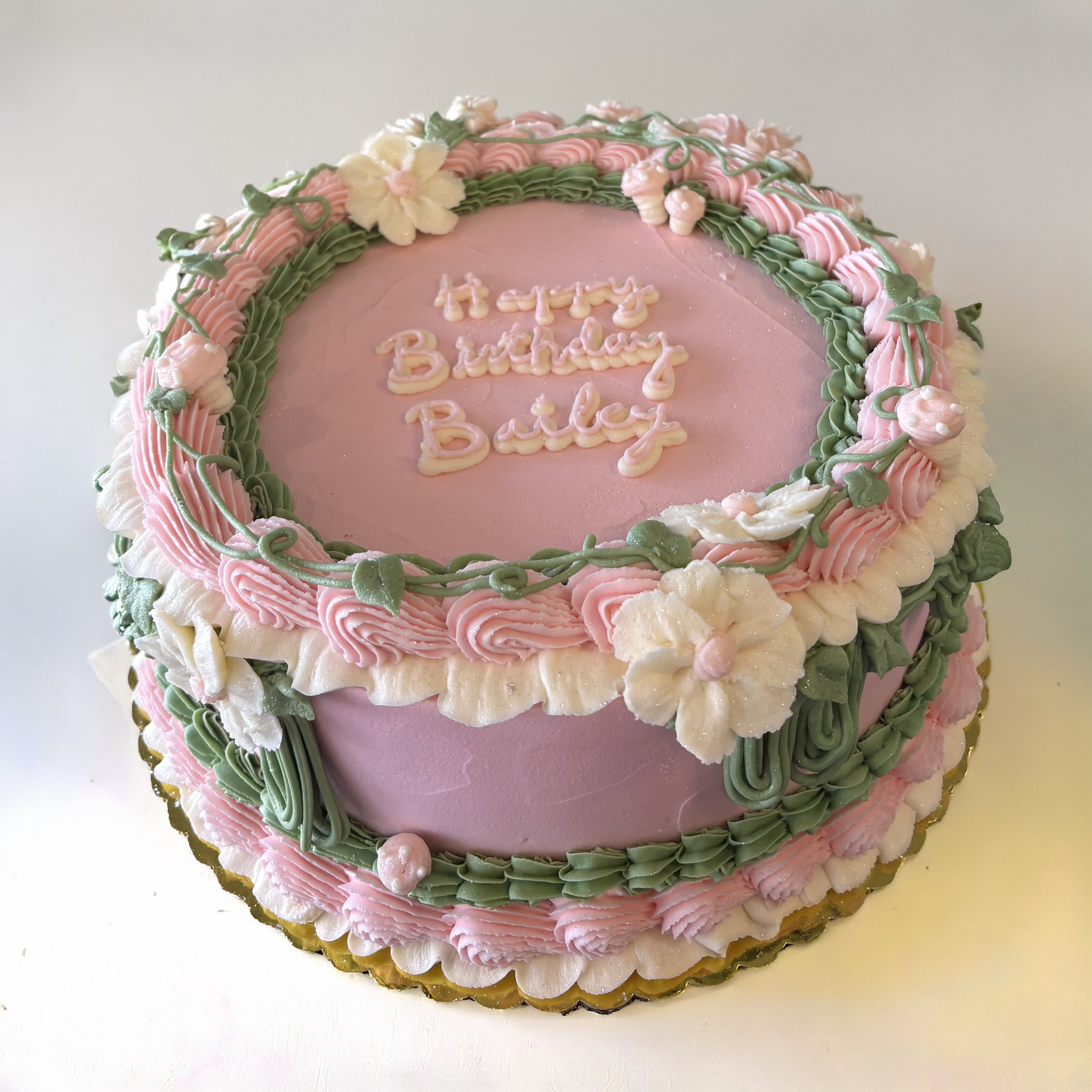 Pink birthday cake decorated with white, pink, and green icing flowers and swirls, with the message 'Happy Birthday Baby Girl' written on top.