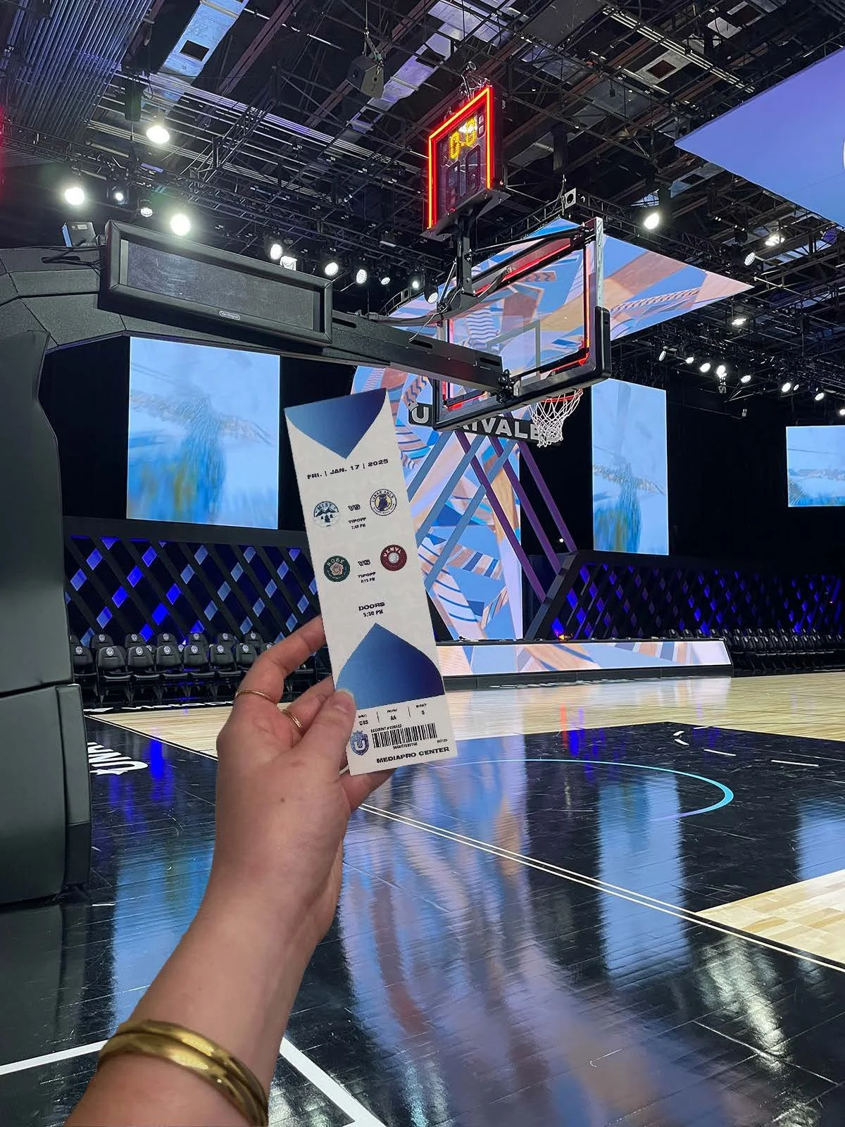 A person holding a ticket for a basketball game at the Mediapro Center, with an empty basketball court and digital screens in the background.