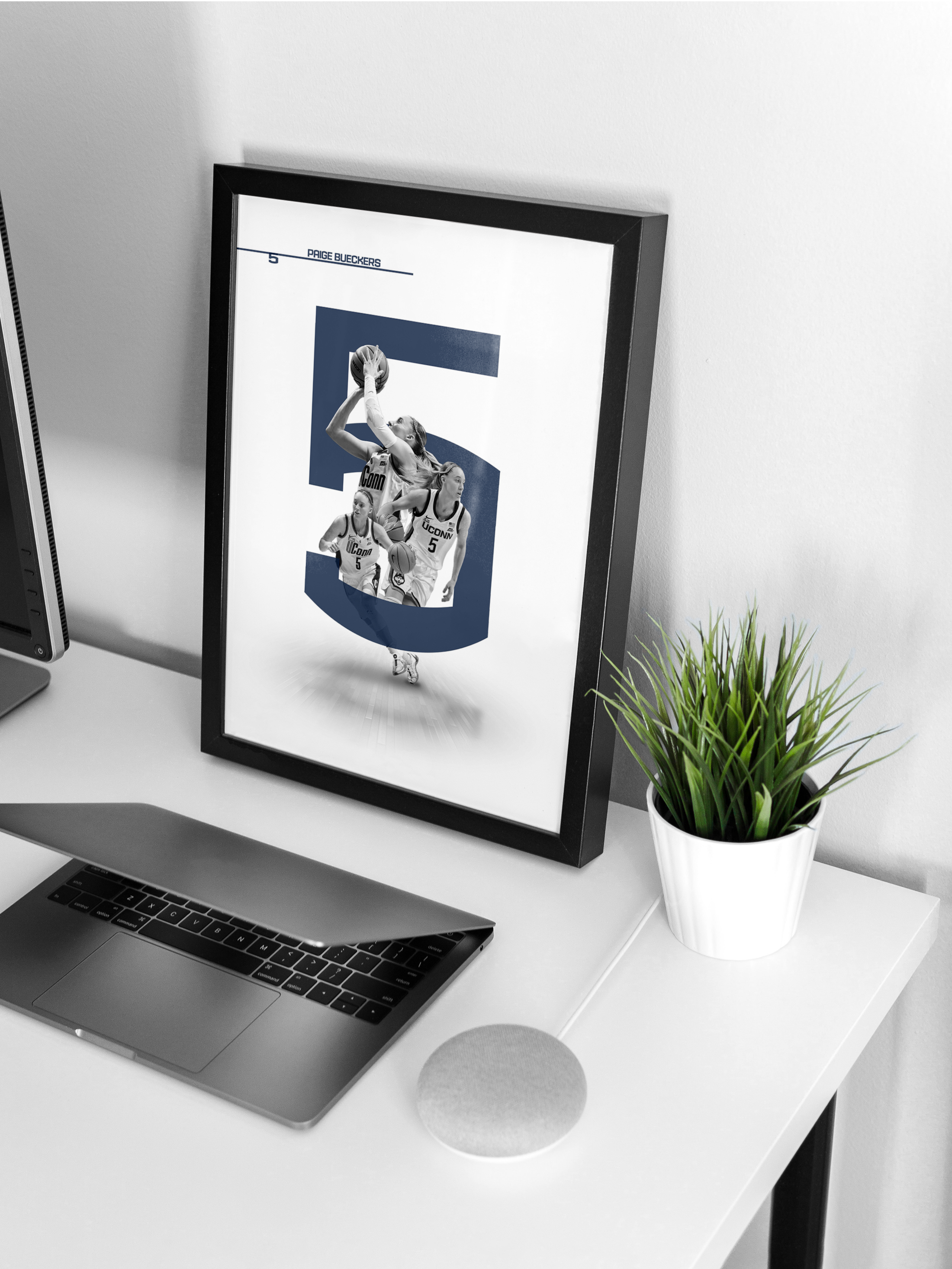 Framed poster on a desk featuring images of women's basketball players with the number 5, against a large number 5 background. The desk also has a laptop, a small speaker, a potted plant, and a monitor.