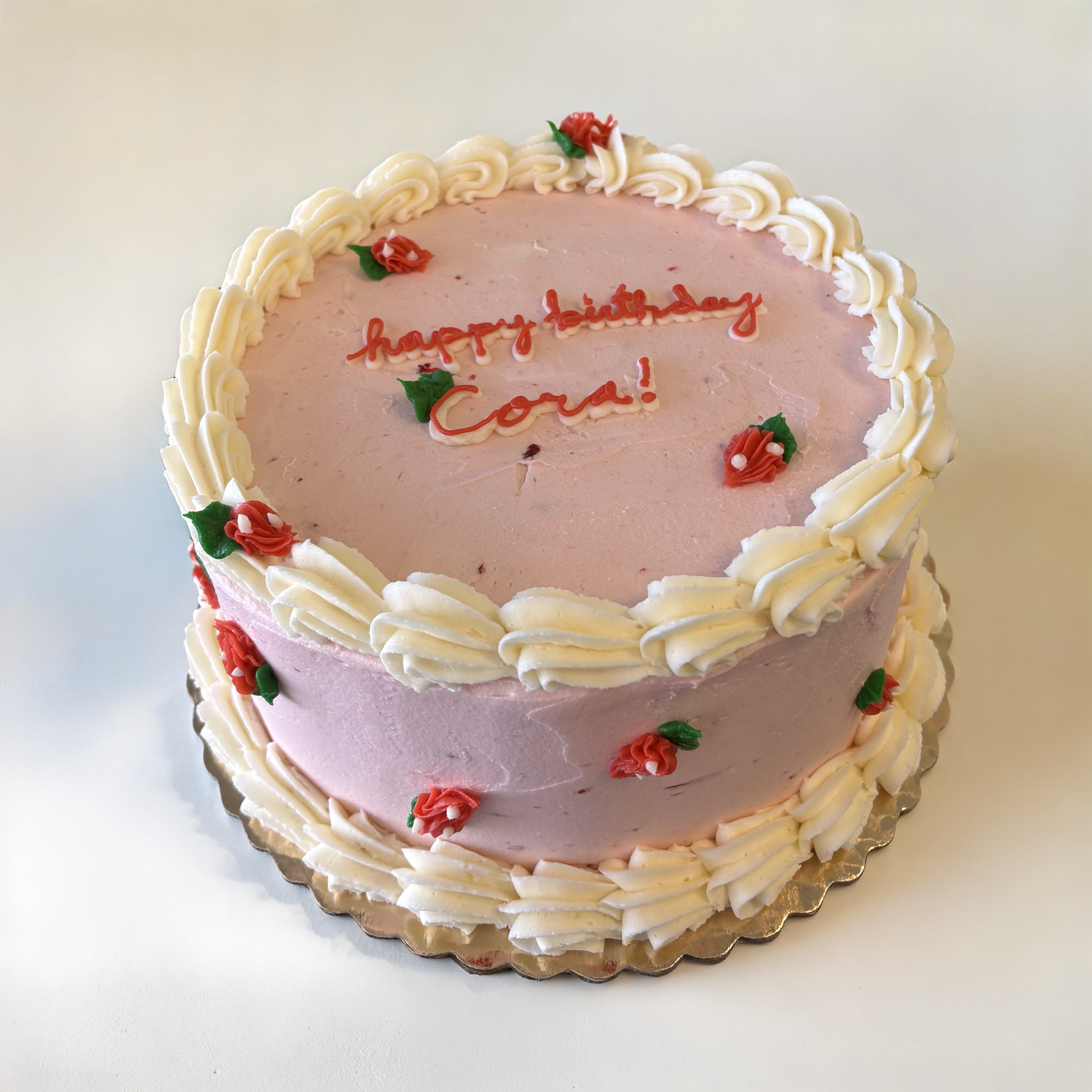 A pink birthday cake with white frosting decorations, small red flowers with green leaves, and the words 'happy birthday Carl!' written in red icing on top.