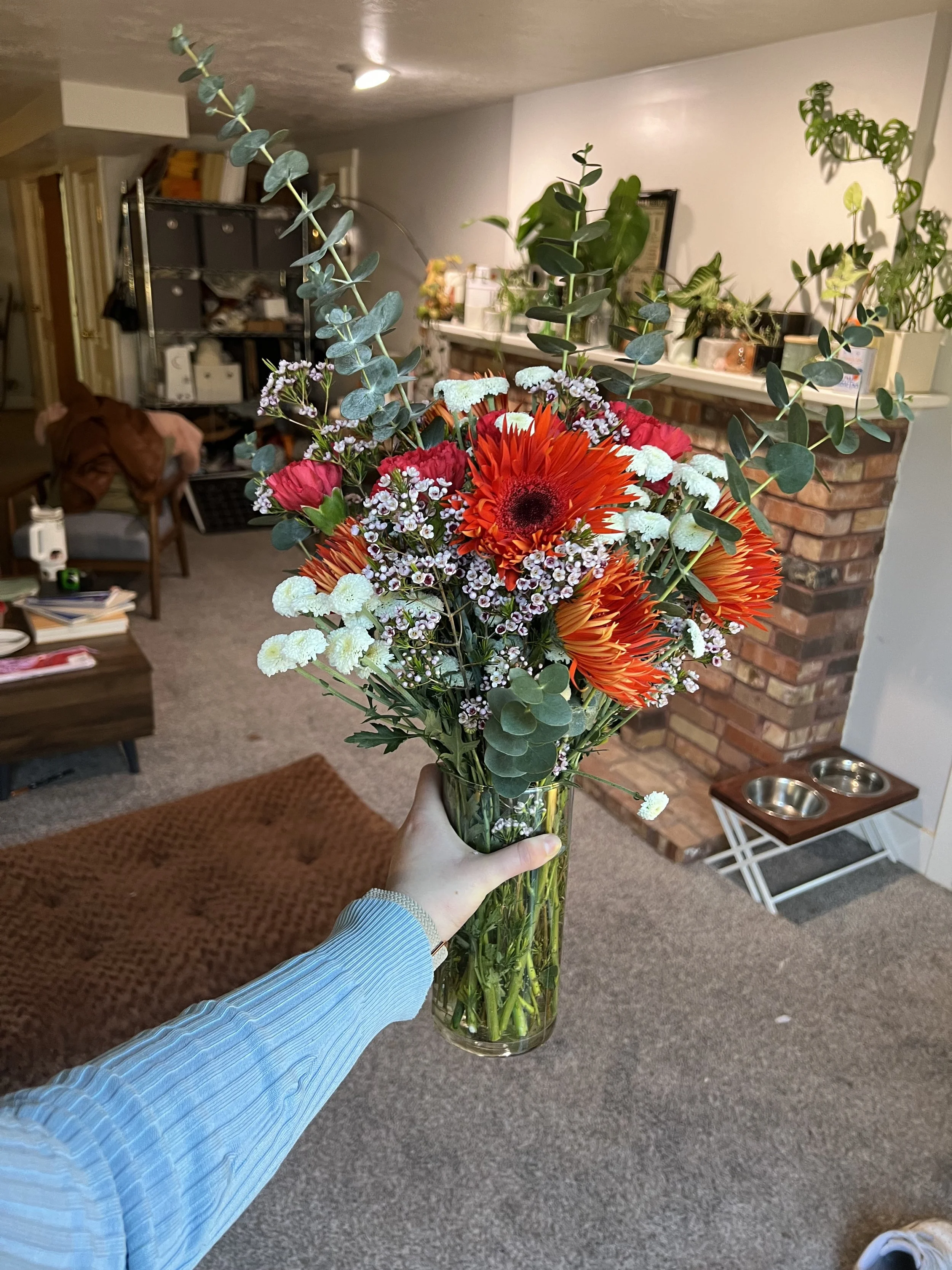 Hand holding a glass vase filled with a bouquet of red, orange, and white flowers and green foliage inside a living room.