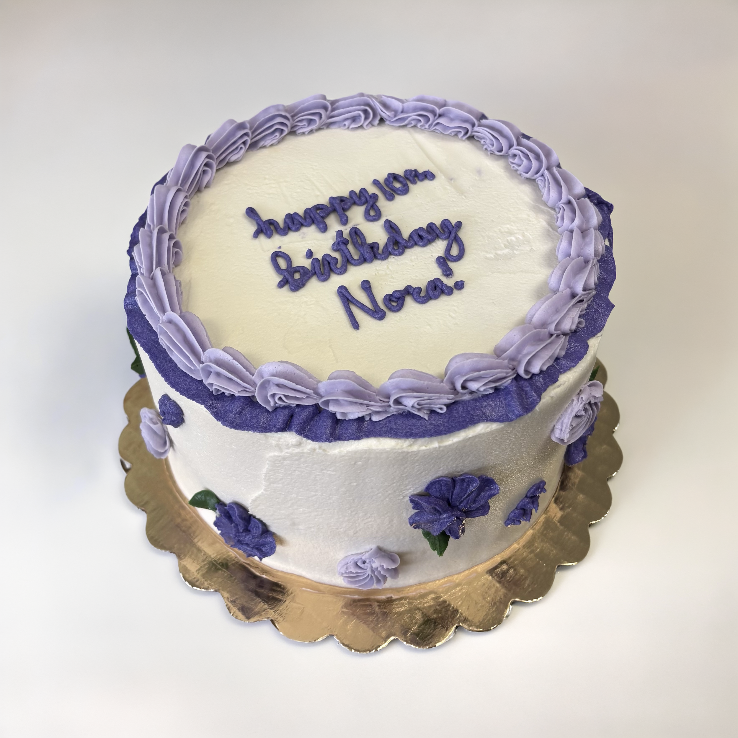 White birthday cake with purple decorative flowers and piping, inscribed with 'happy 10th Birthday Nora' in purple icing, on a gold scalloped cake board.
