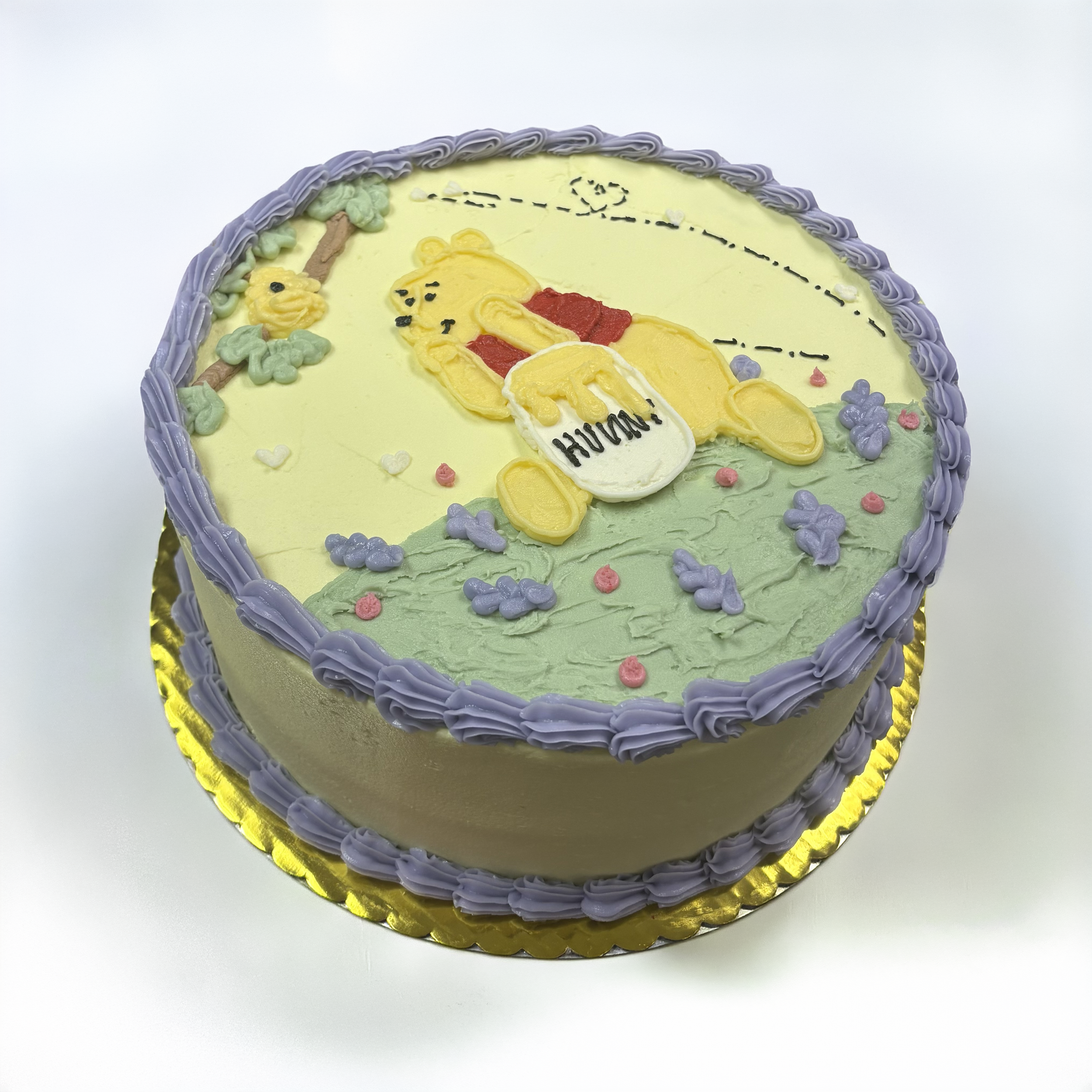 A decorated round birthday cake with purple icing border, yellow and green icing details, and a Winnie the Pooh themed design featuring Pooh, a honey jar, and the words 'Happy Birthday' written on the cake.