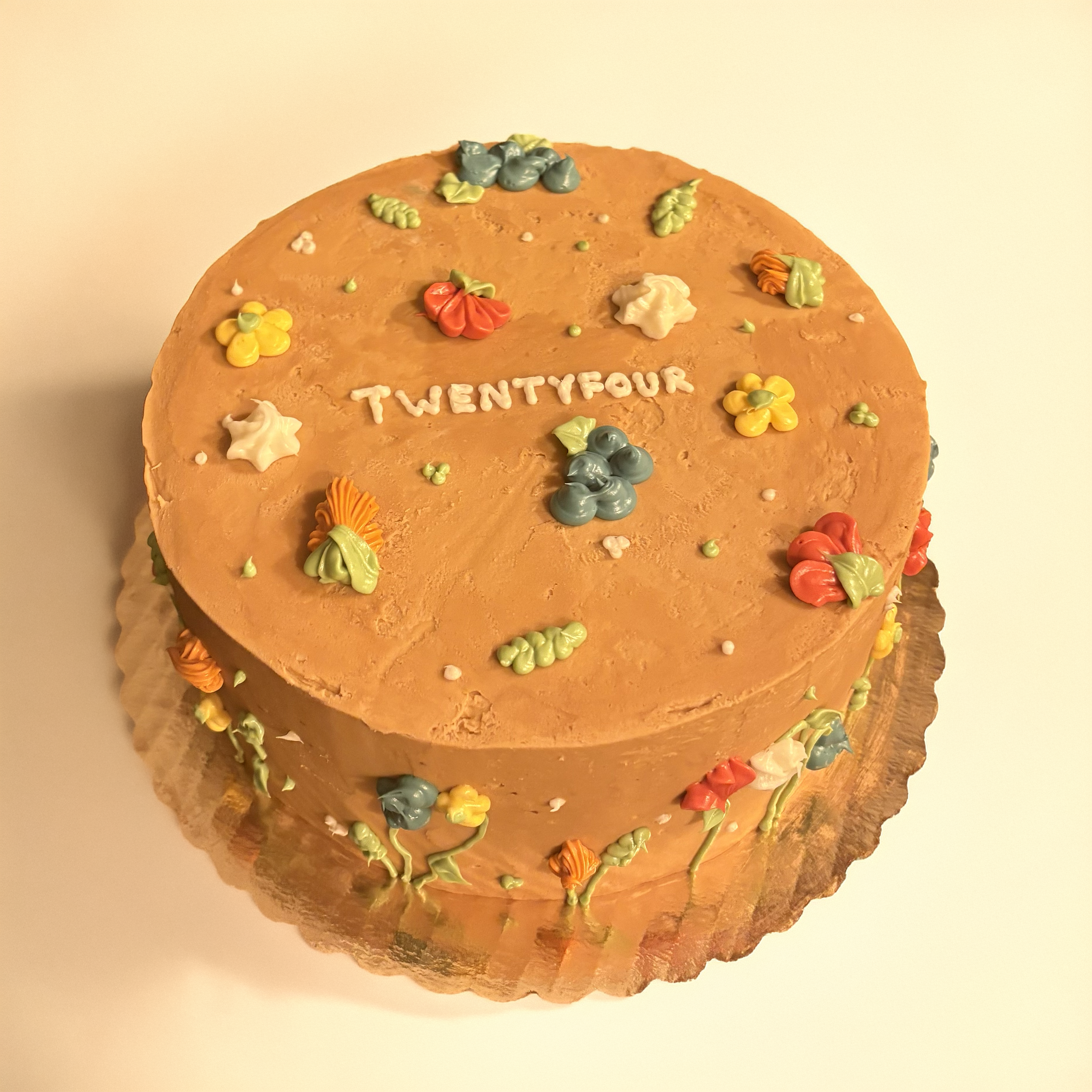 Round birthday cake with orange frosting, decorated with colorful flowers made of icing, and the number twenty-four written in white icing on the top.