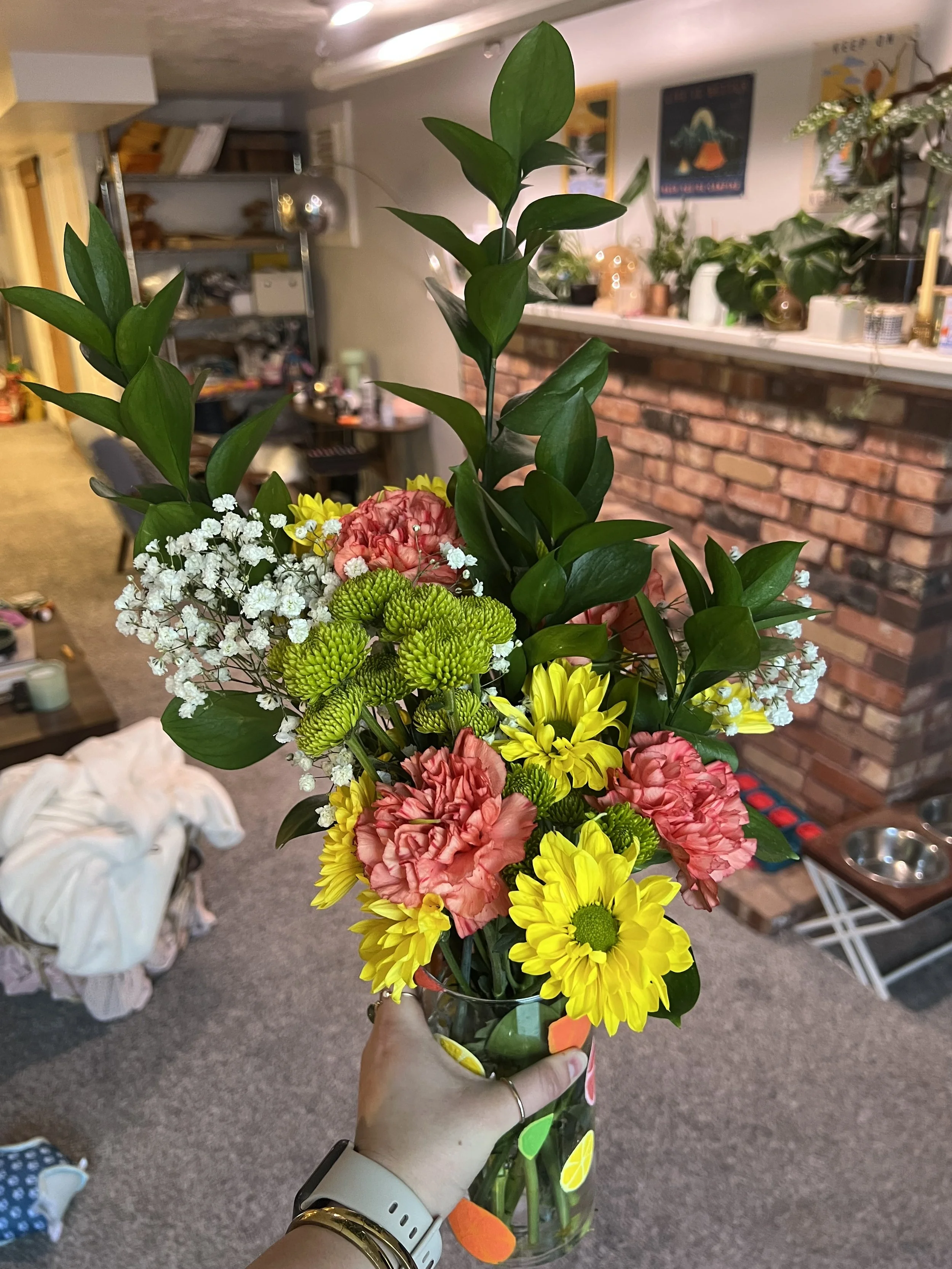 A person holding a colorful bouquet of flowers in a glass vase indoors, with a brick fireplace and various household items in the background.
