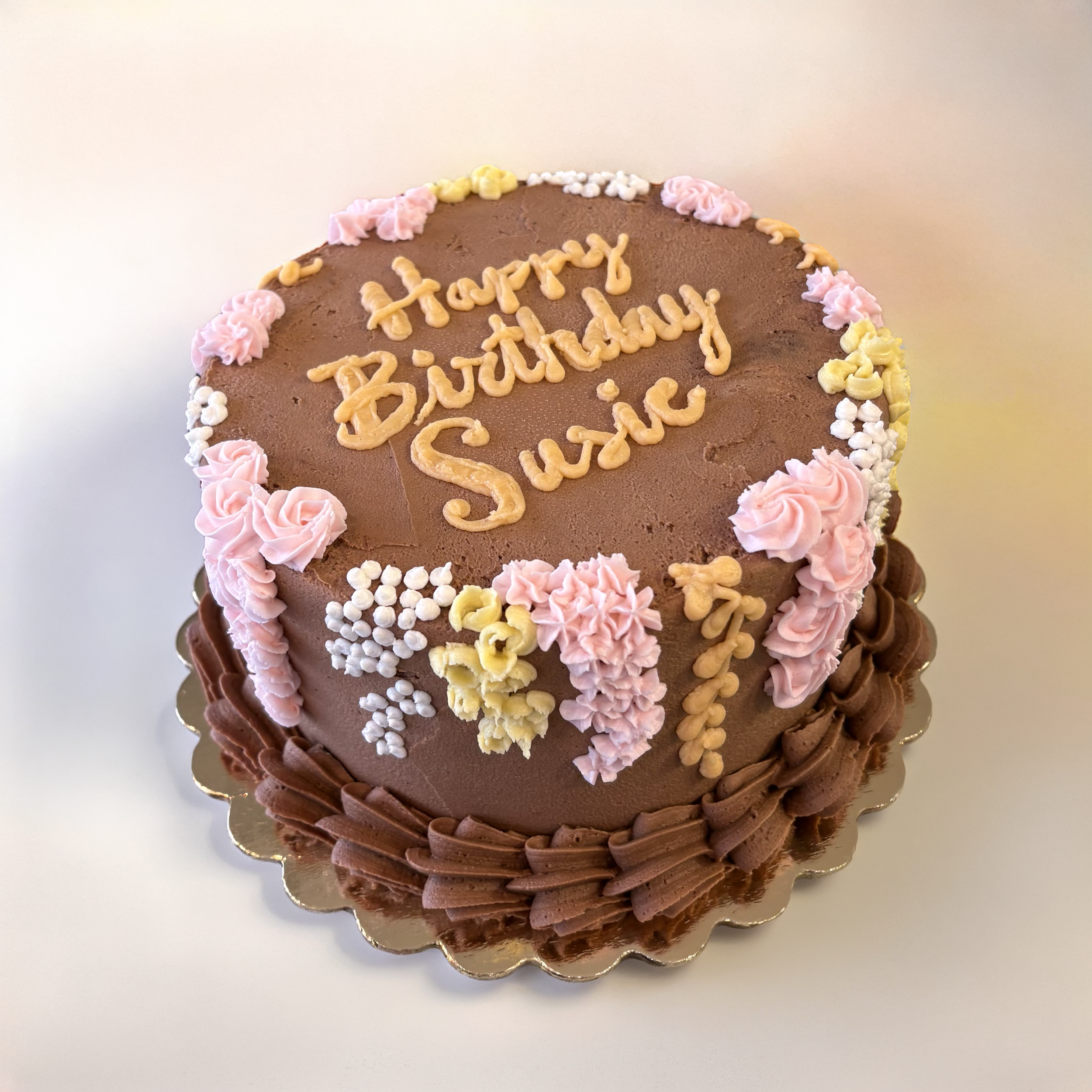 Chocolate birthday cake decorated with pink, yellow, and white buttercream flowers with a message written in yellow icing on top.