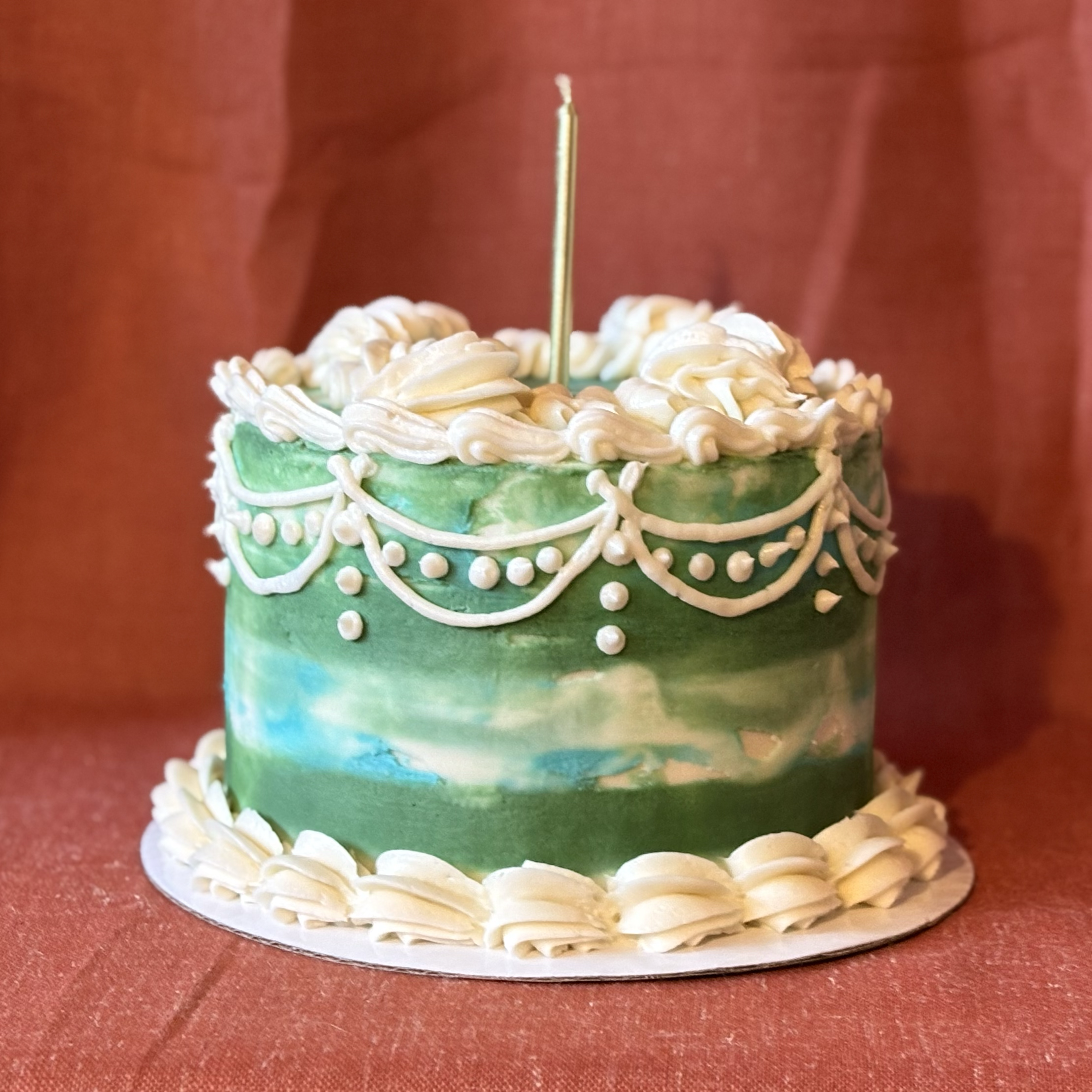 A decorated birthday cake with green watercolor-like icing, white piped frosting at the top and bottom, and white decorative piping near the top. A single tall candle is inserted in the center.