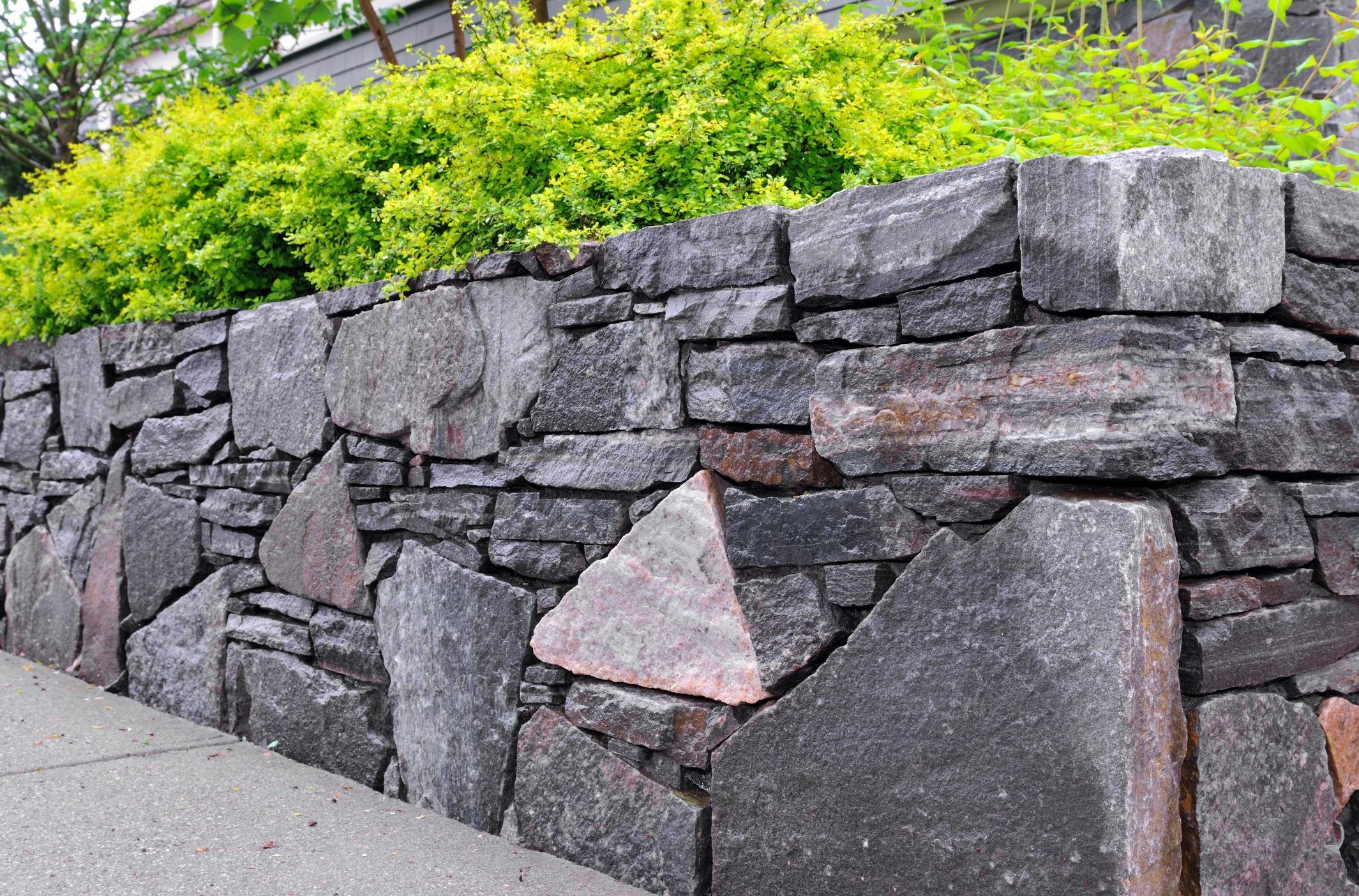 Stone retaining wall with green shrubbery behind it and part of a sidewalk in front.