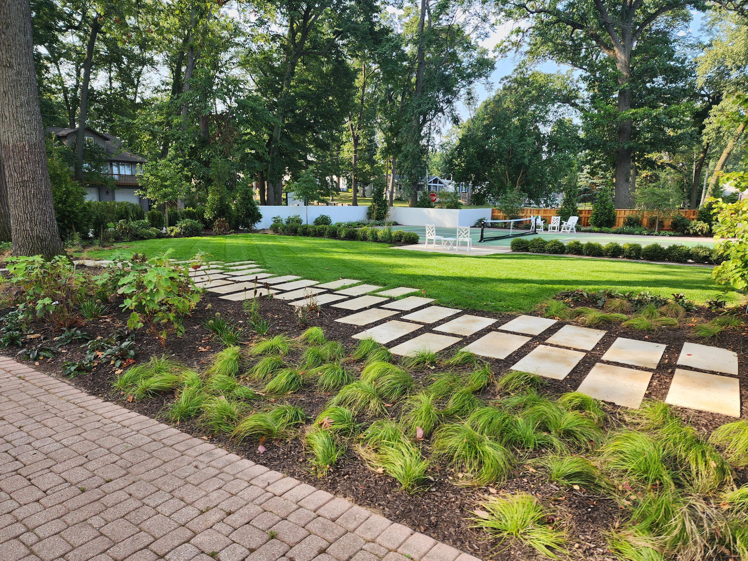 A backyard with a stone pathway, green grass, garden beds, trees, and white outdoor furniture.