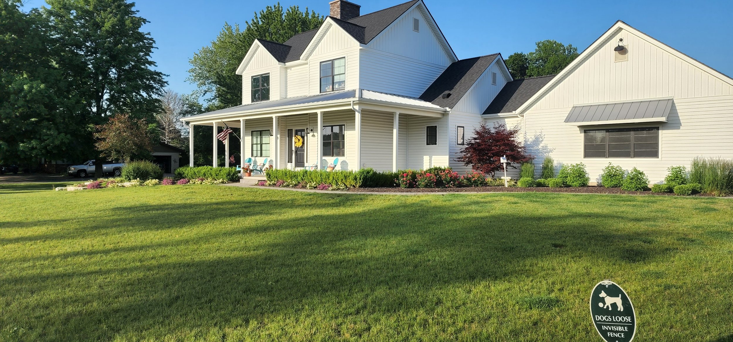 White two-story house with a front porch, surrounded by a green lawn and landscaped garden with bushes and trees, and a sign that says "Dogs Loose Invisible Fence" in the yard.