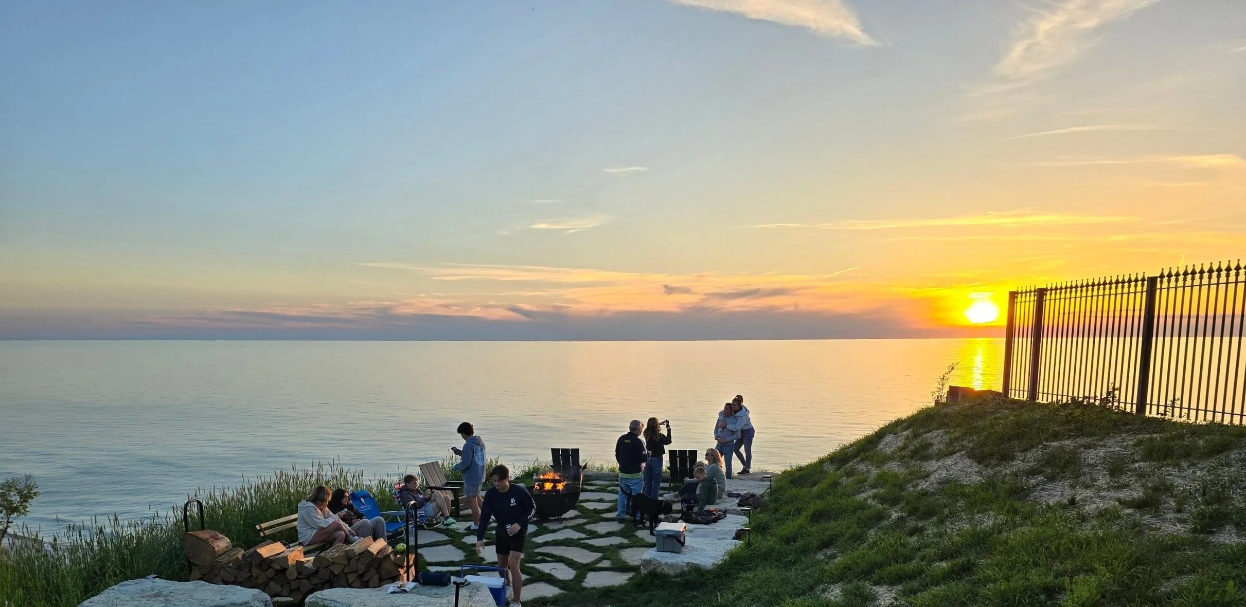 Group of people gathered near a fire pit on a seaside patio during sunset, with the ocean and colorful sky in the background. Some are sitting, some standing, and a couple is hugging near the fence on the hillside.