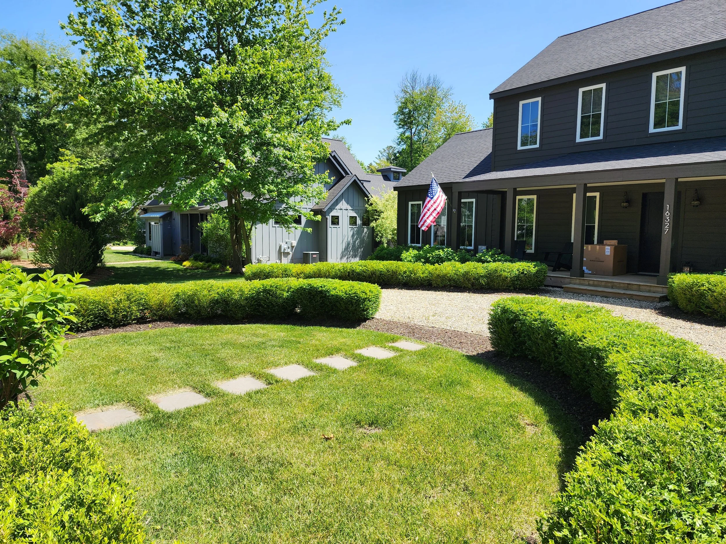 A well-maintained front yard with green grass, trimmed bushes, and a large tree next to a black house with a covered porch. An American flag is hanging on the porch. There are stepping stones on the grass and a gravel pathway leading to the porch.