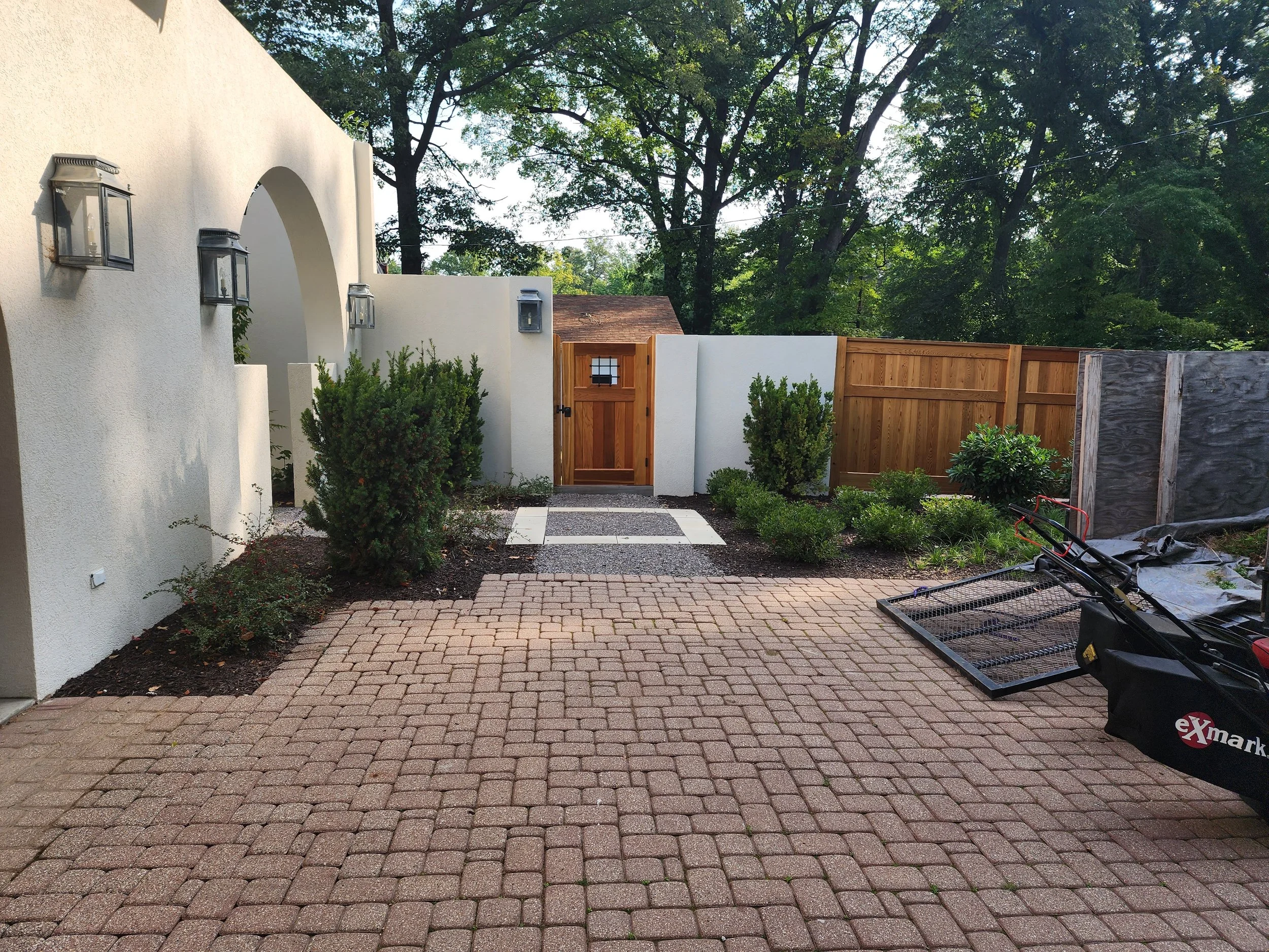 A backyard patio with a brick paver surface, a white stucco wall with outdoor lanterns, a small wooden gate, green shrubs, and a wooden fence, with construction or gardening equipment on the right side.