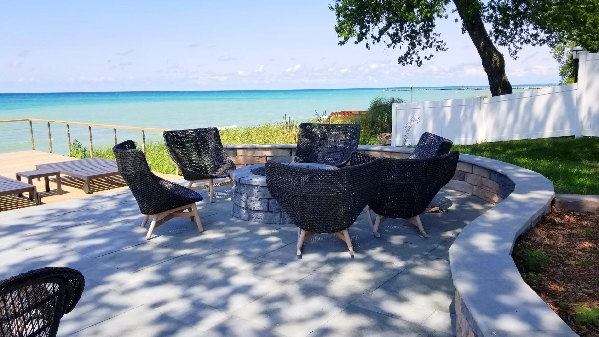 Beachfront patio with black chairs and fire pit, overlooking ocean with grassy area and trees.