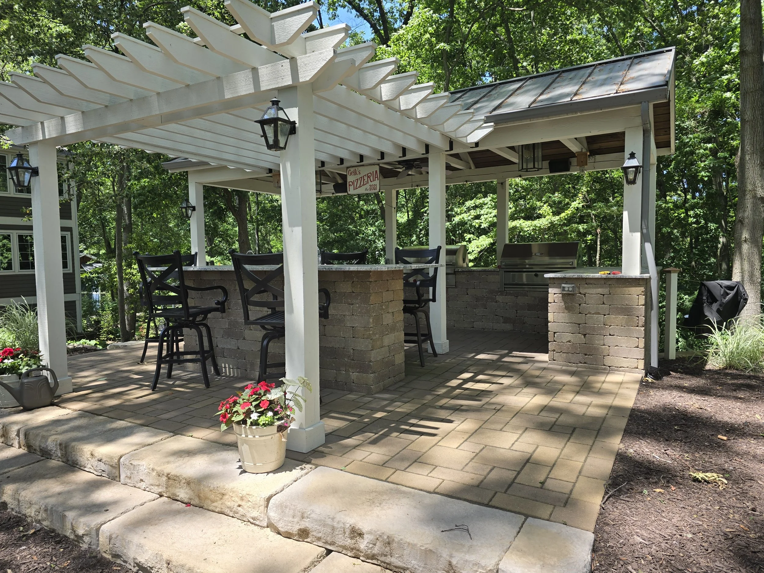Outdoor kitchen and bar area with a pergola, brick countertops, bar stools, a grill, and a sign for a pizzeria, surrounded by trees and greenery.
