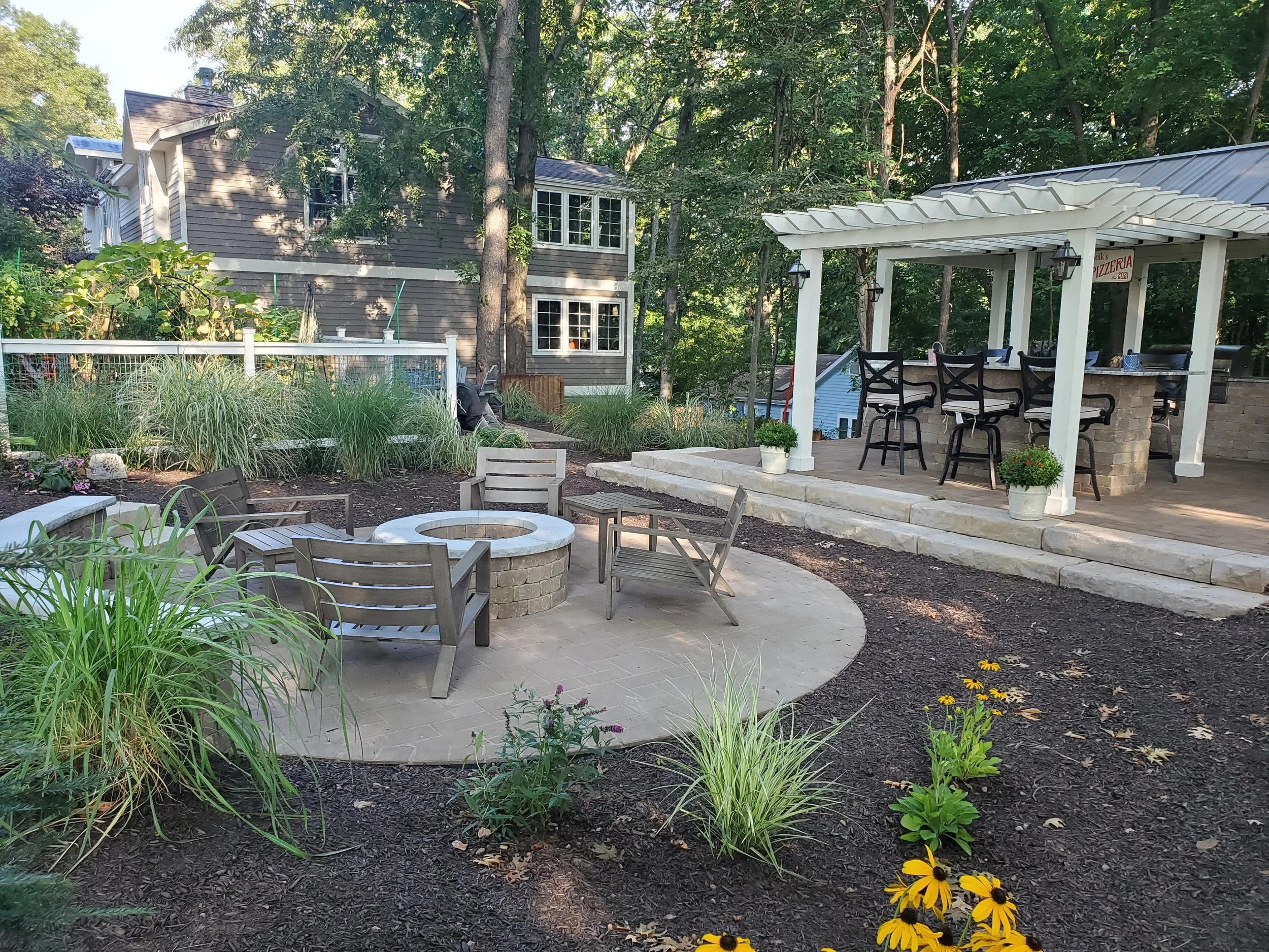 A backyard patio area with outdoor seating, a fire pit, and a covered bar area. The yard features plants, flowers, and trees with a house in the background.