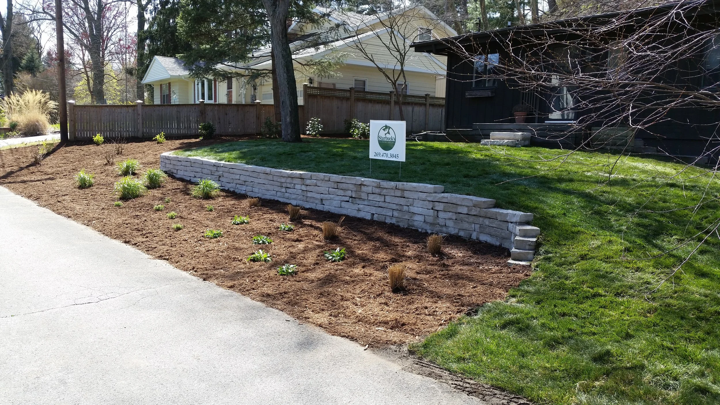 A landscaped front yard with a gradient brick retaining wall, freshly planted small plants, and a lawn. There is a sign with a green leaf logo and text in front of the lawn.