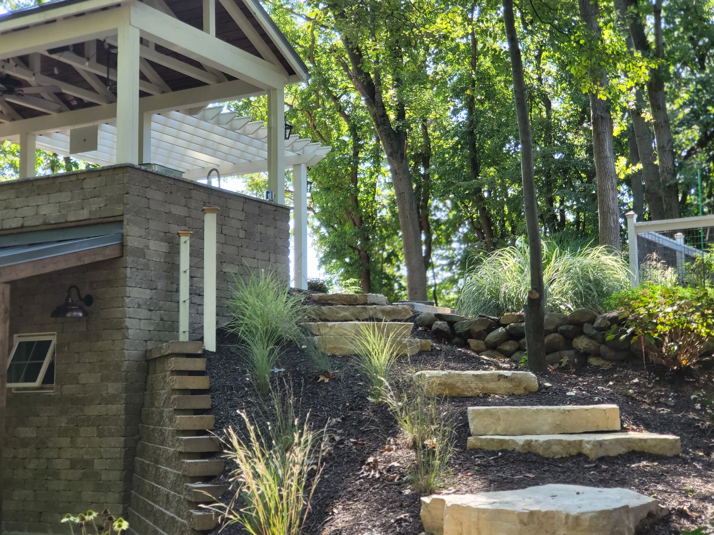 Stone stairs leading up a slope with plants and trees surrounding a backyard patio with a white pergola.