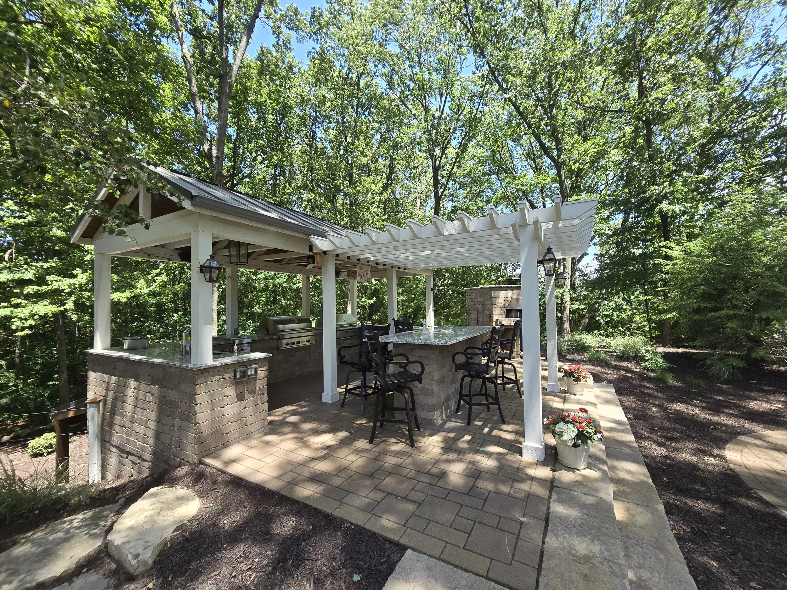 Outdoor covered kitchen and bar area with grill, bar stools, potted flowers, surrounded by trees.