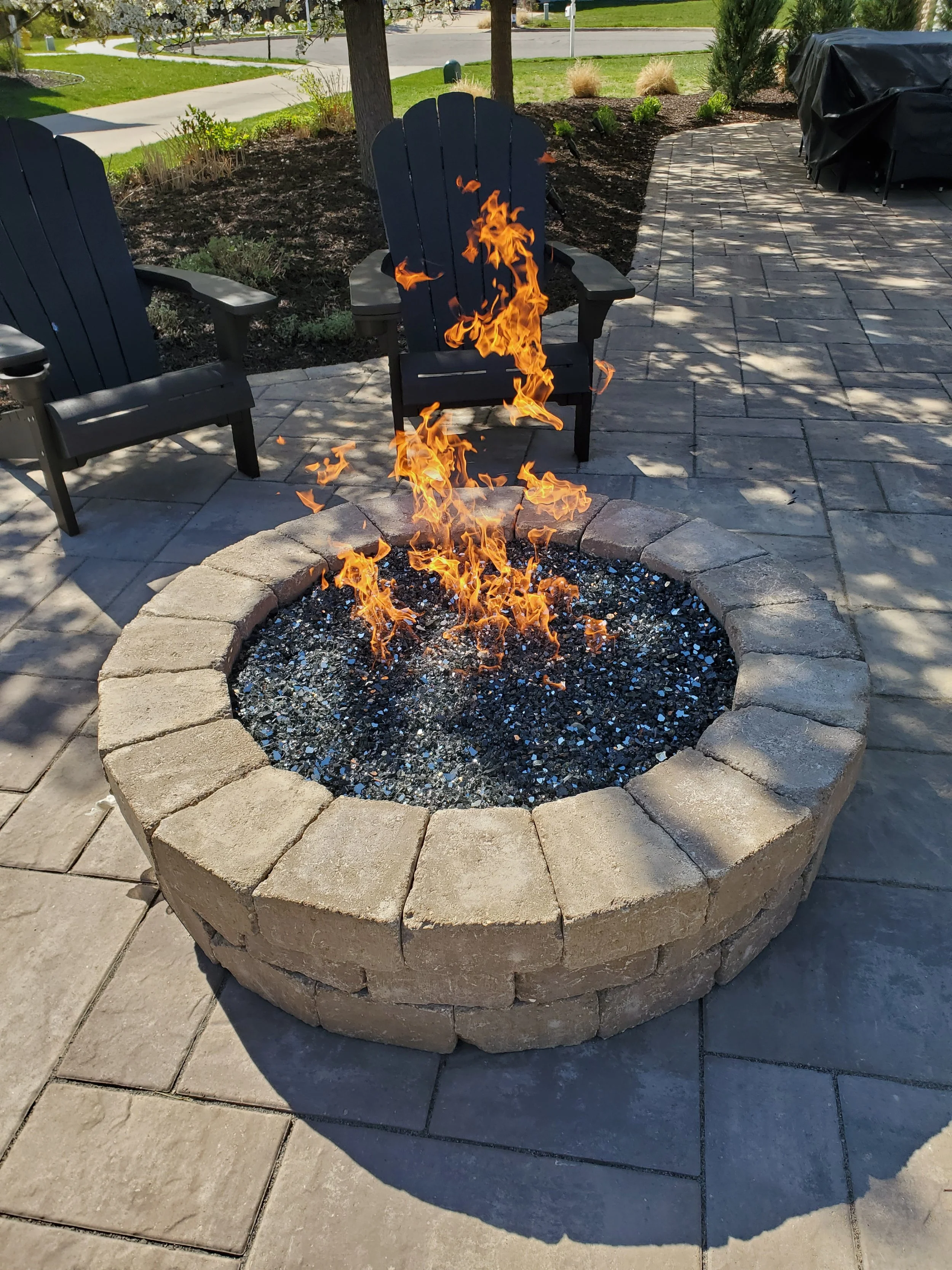 A circular fire pit made of beige bricks with flames rising from black gravel inside, surrounded by a paved patio with two black Adirondack chairs in the background.
