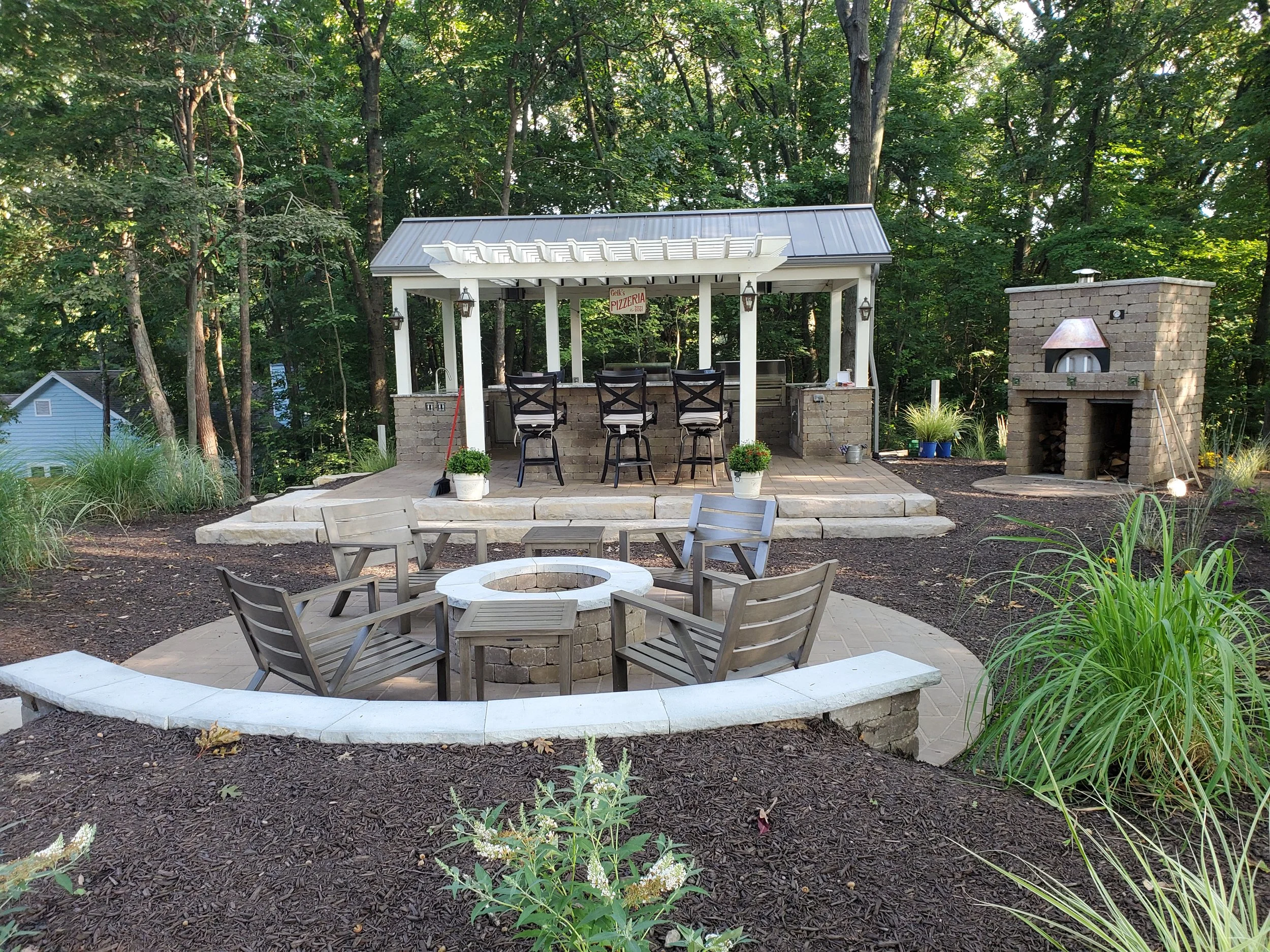 Outdoor backyard with a stone fire pit surrounded by wooden chairs, a covered bar area with stools, and a brick outdoor oven, all set amidst landscaped garden with trees and plants.
